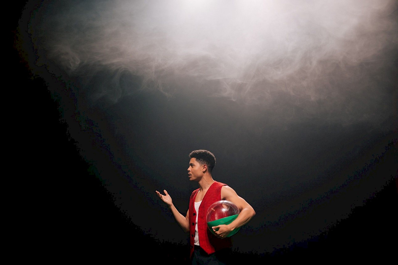 ALKANARA - Marco Mendonça, de colete vermelho, segura num globo insuflável e colorido enquanto gesticula expressivamente contra um cenário escuro e cheio de fumo. - ©Diana Tinoco