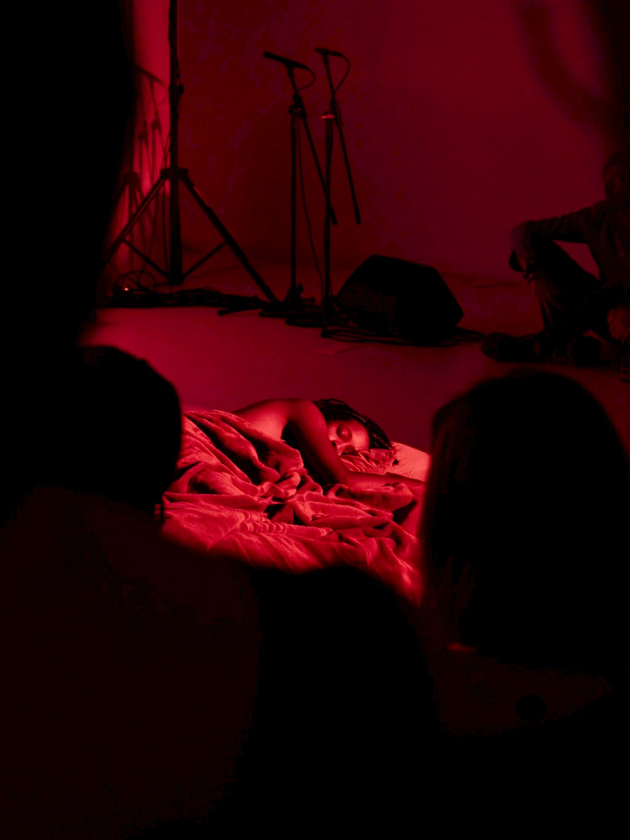 ALKANARA - Uma pessoa deitada num cobertor, dormindo pacificamente sob luz vermelha, com membros do público visíveis em primeiro plano, observando a cena. - ©Fabiana Tavares