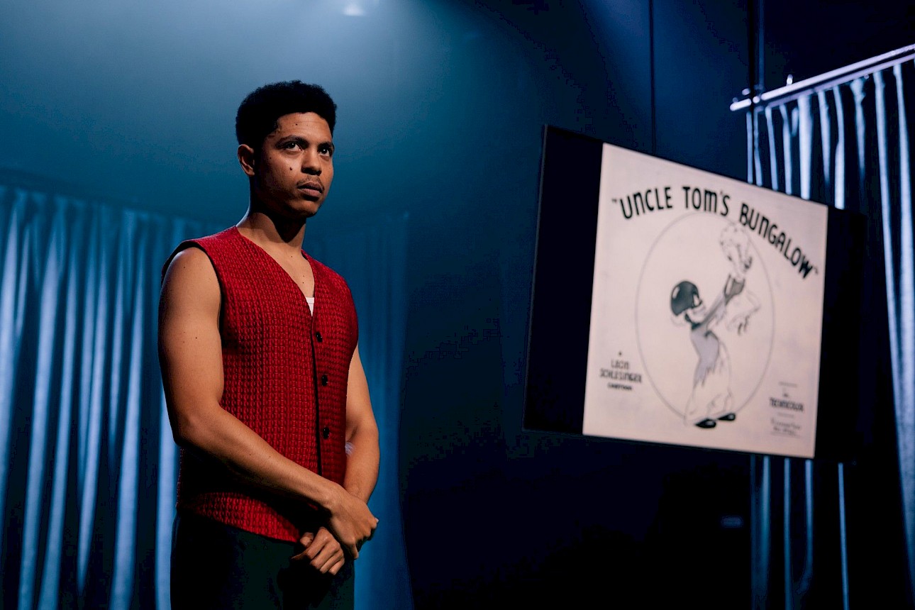 ALKANARA - On the left side of the photograph, Marco Mendonça appears in the foreground, wearing a red waistcoat. He's standing, his hands are crossed in front of his body and he's looking straight ahead. On the right, out of focus, is a suspended screen showing a black and white image with no definition. In the background are long curtains, coloured blue by the projection of the lights - ©Diana Tinoco