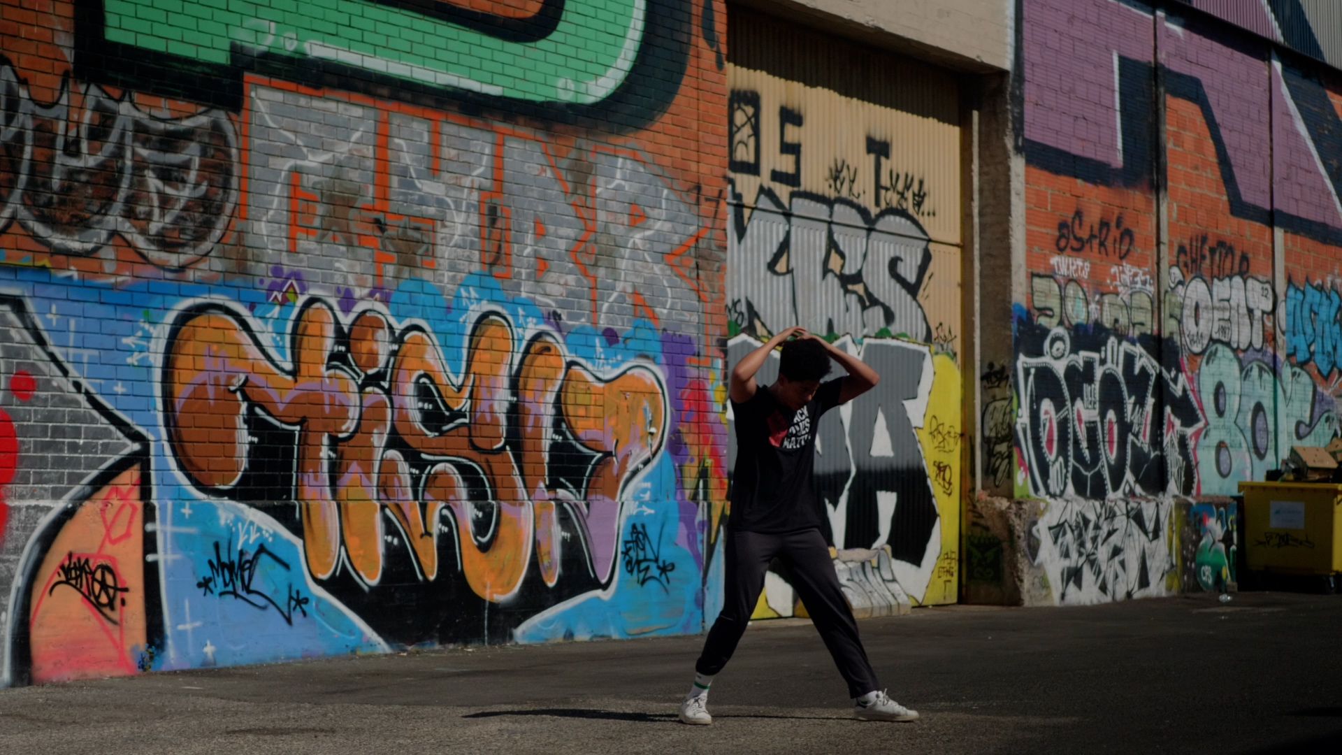 Uma pessoa está de pé, com os braços no ar, em frente a um muro de graffiti colorido. - Heverton Harieno