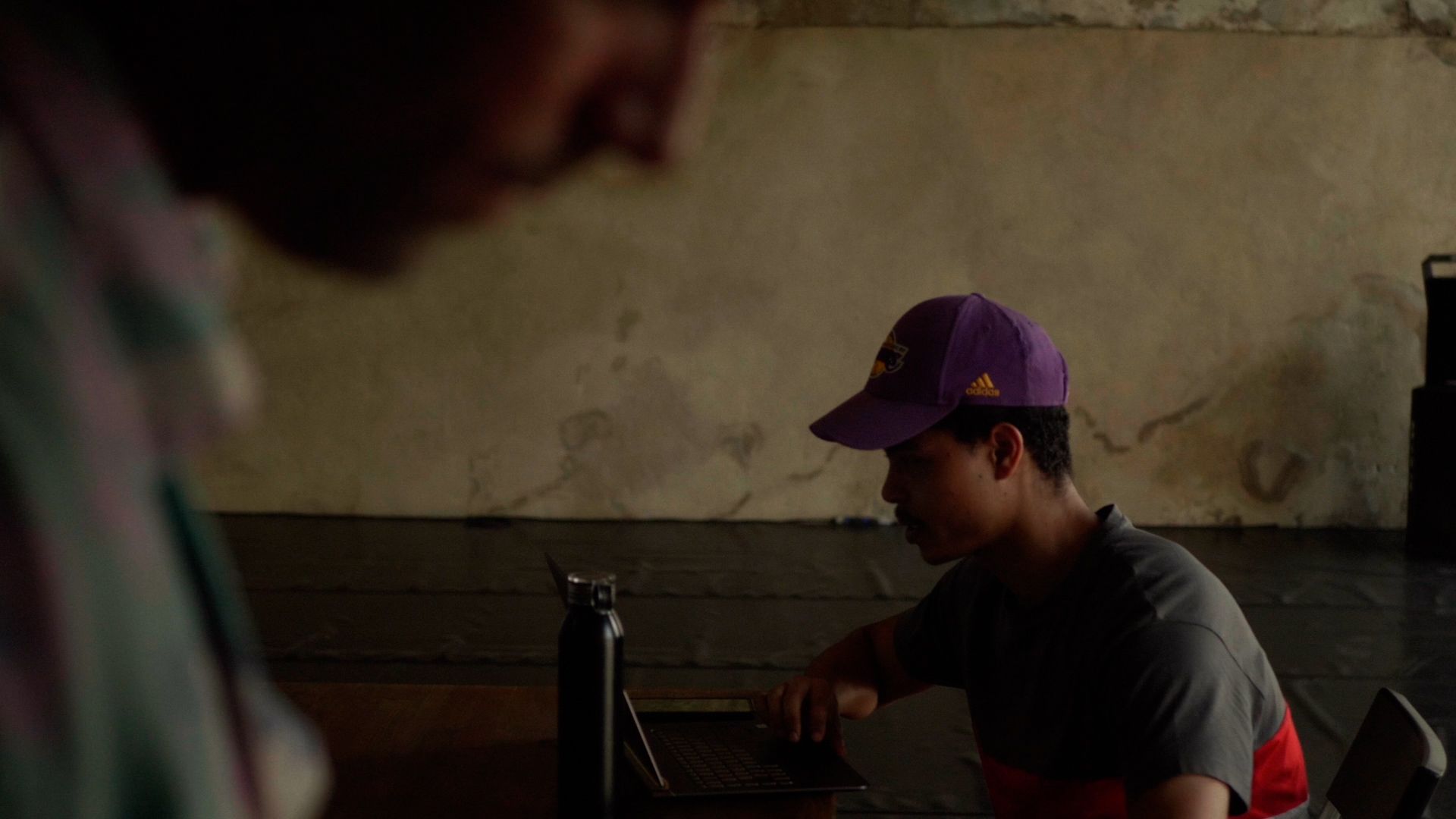 Pessoa com um boné roxo a utilizar um computador portátil numa mesa com uma garrafa de água. - Heverton Harieno