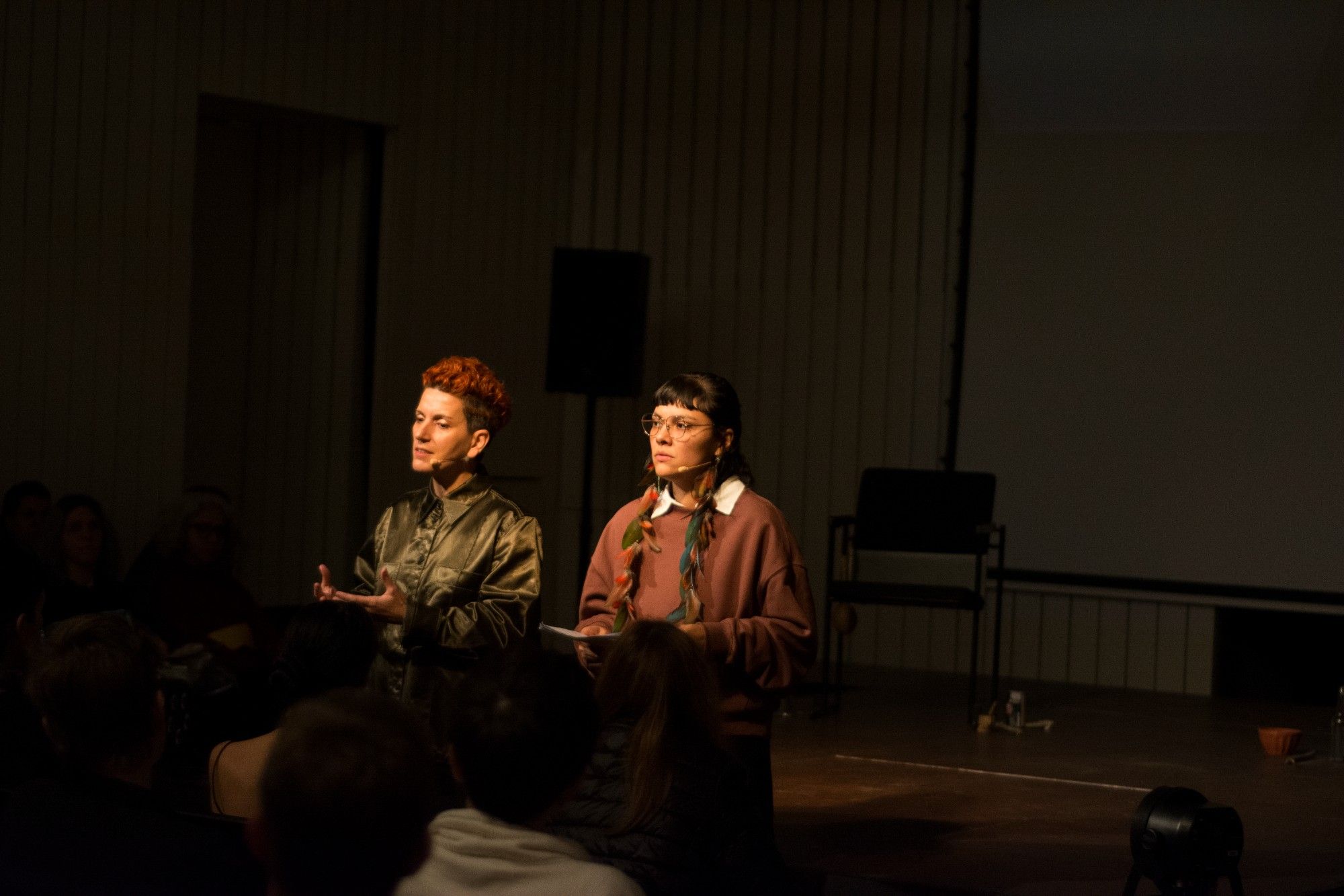 Two individuals standing on stage addressing an audience in a dimly lit room. - Rui Palma