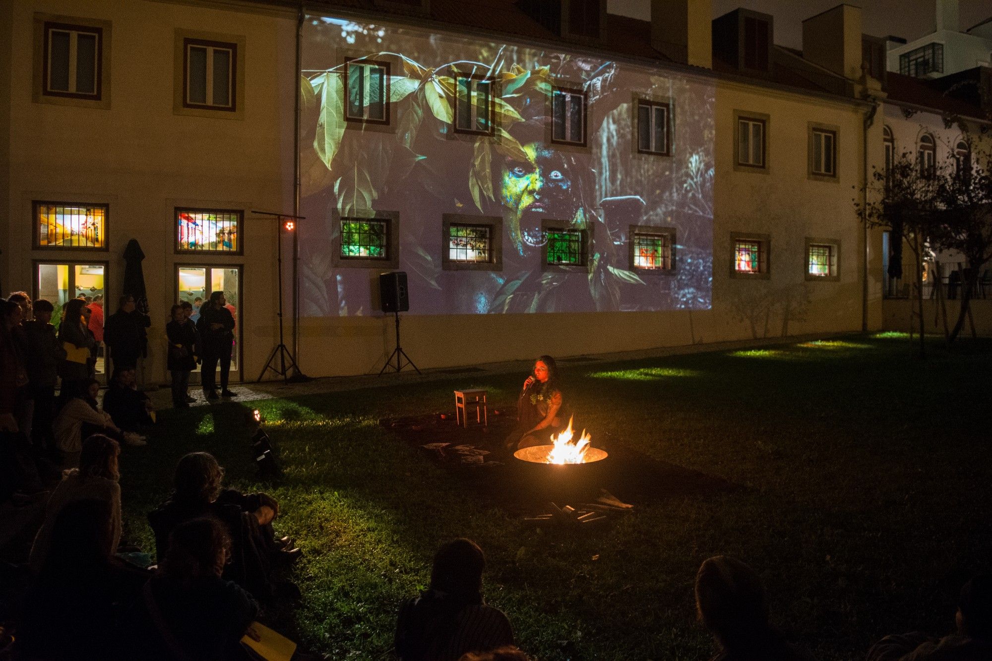 Projeção ao ar livre num edifício com uma audiência em volta de uma fogueira à noite. - Rui Palma