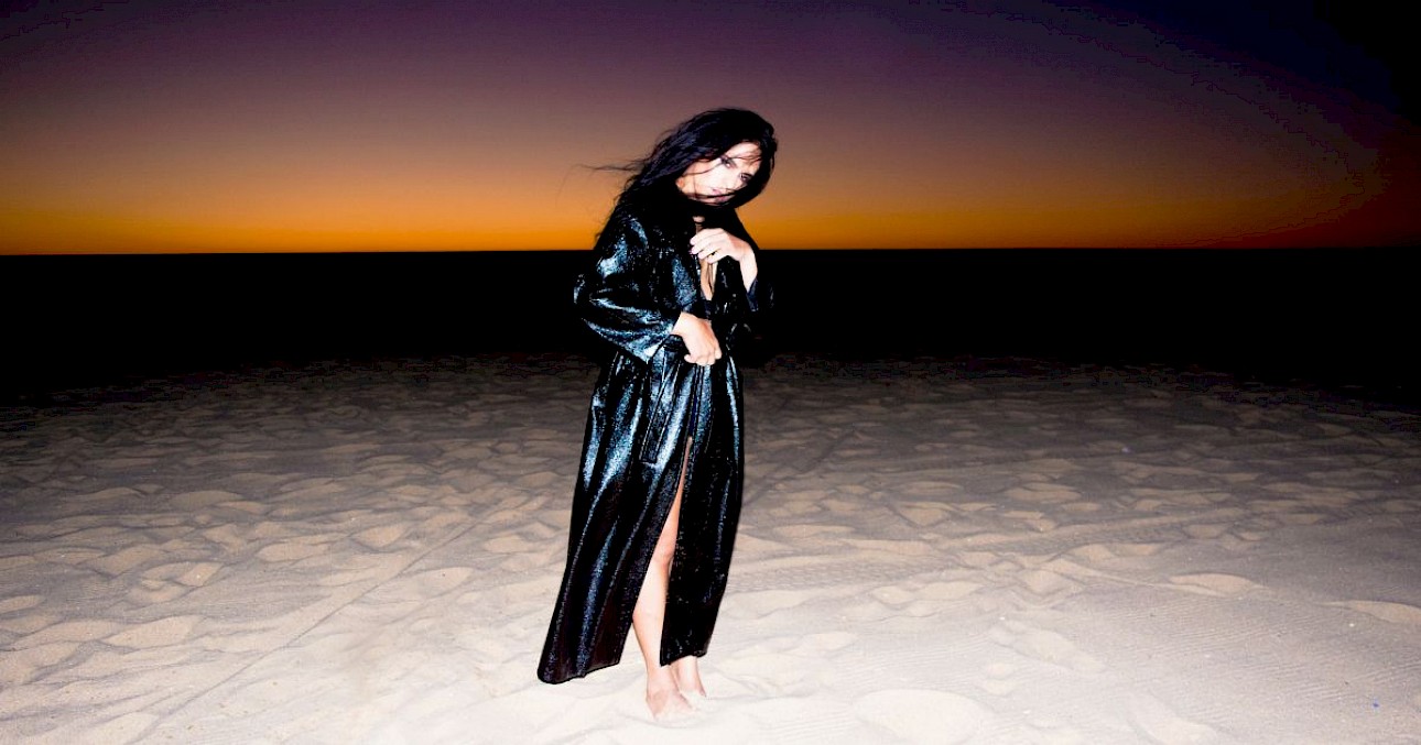 ALKANARA - Diego Bragà is in the center of the photograph, looking directly into the lens, with arms hanging by their side. They are dressed in a long, shiny black coat, reaching down to their bare feet buried in the sand. In the background, the horizon line and the evening sky, fading from orange to purple. - ©Rui Palma