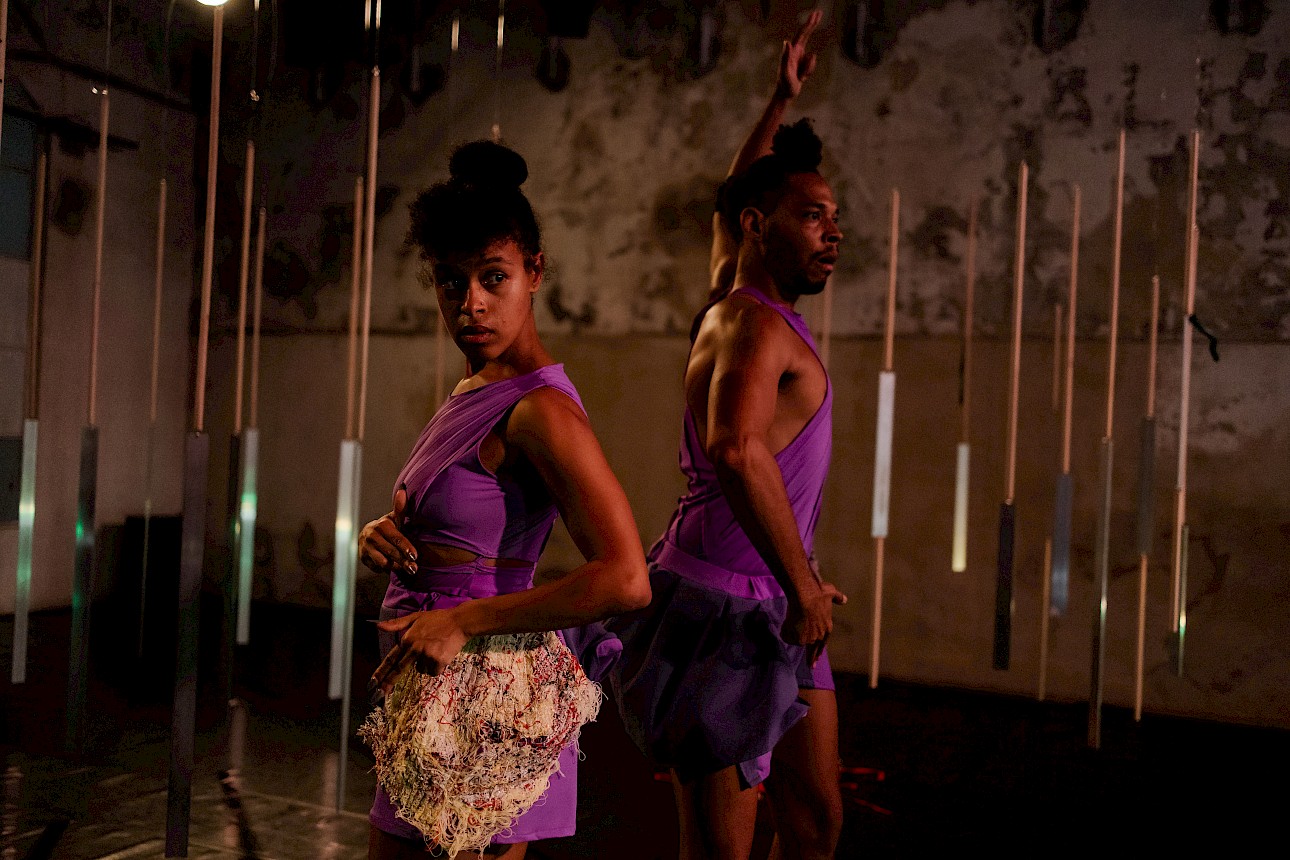 ALKANARA - Two people stretching on a shiny floor, one in a jersey and the other in patterned clothing, with heads down and arms extended forward. - ©Letícia Diniz