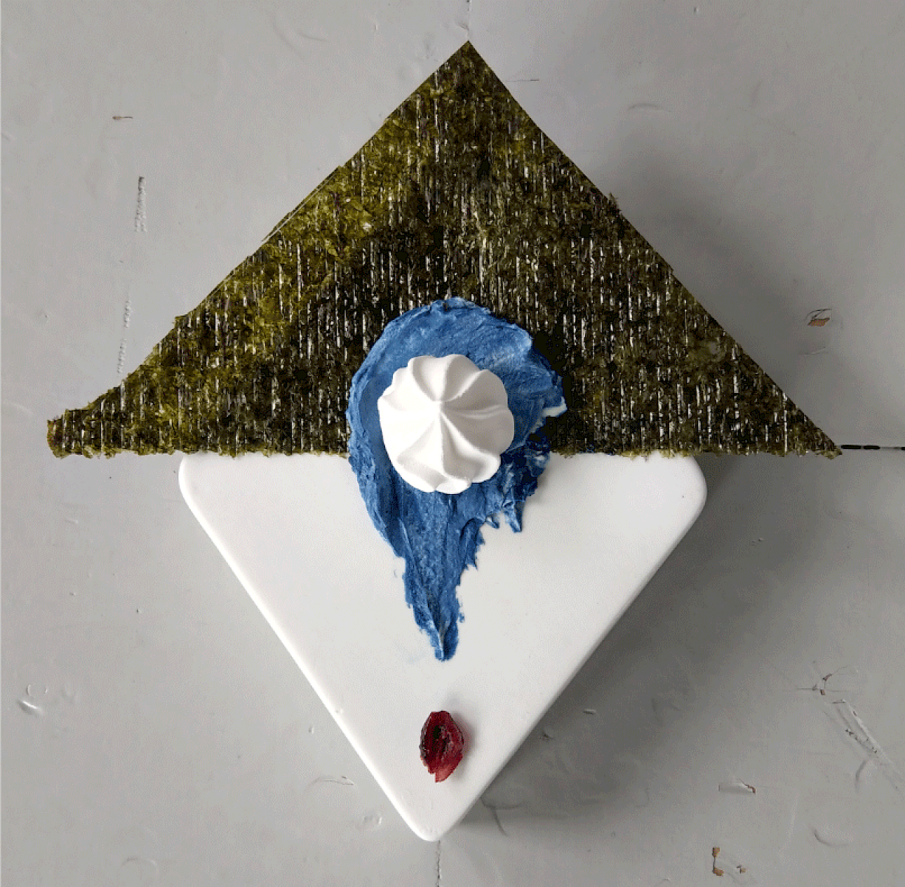 ALKANARA - An artistic dessert with a triangular green topping, blue frosting, a whipped cream dollop, and a red berry on a white plate. - ©Celestial Bodies