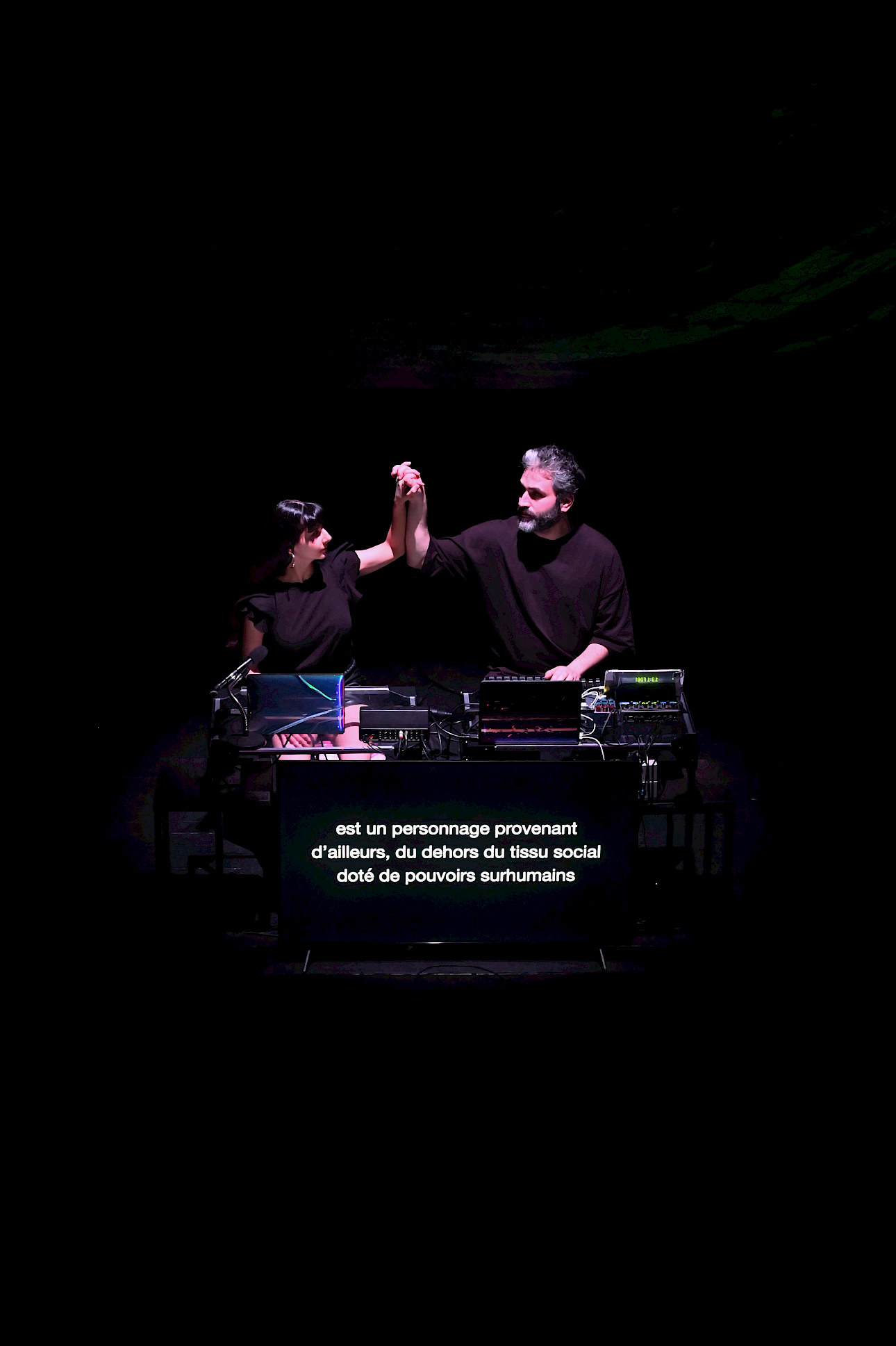 ALKANARA - Two individuals perform on stage, raising their hands together amidst a backdrop of low lights and electronic equipment. - ©Margot Briand