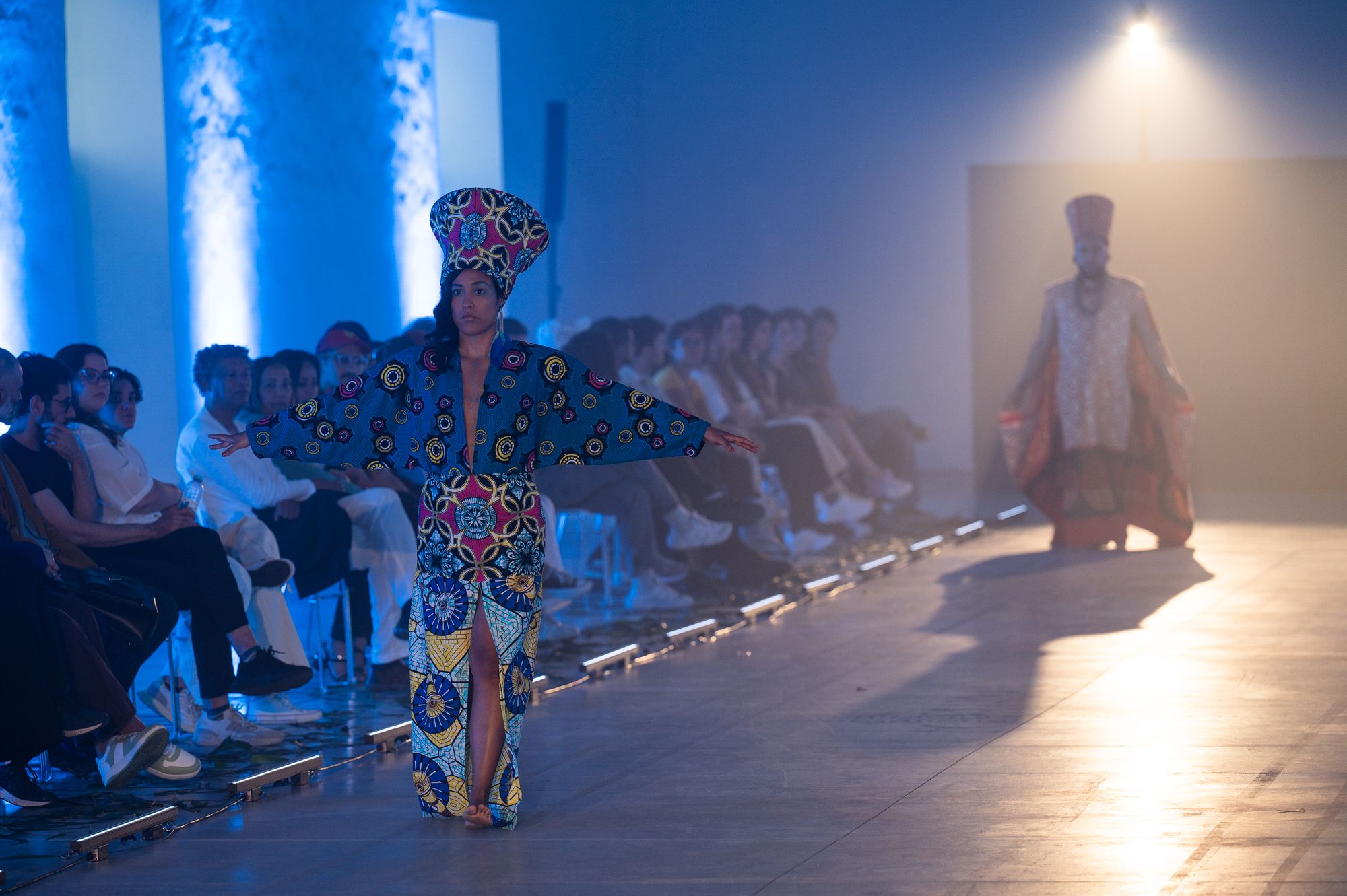 A model showcases vibrant, patterned attire on a runway, with an audience seated in a warmly lit venue. - José Caldeira