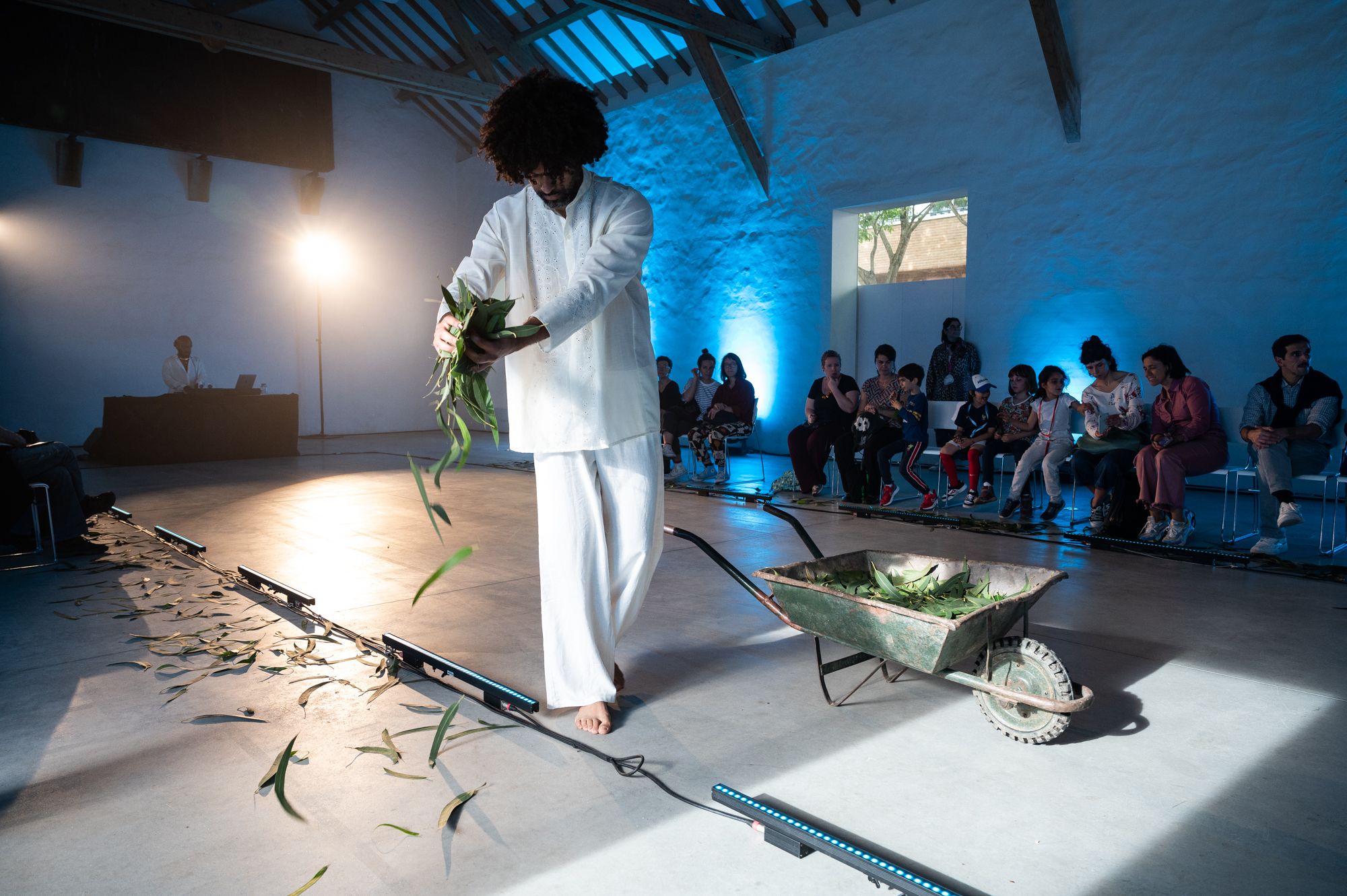 A performer in white attire walks barefoot, holding greenery, while an audience observes in a dimly lit space with blue accents. - José Caldeira