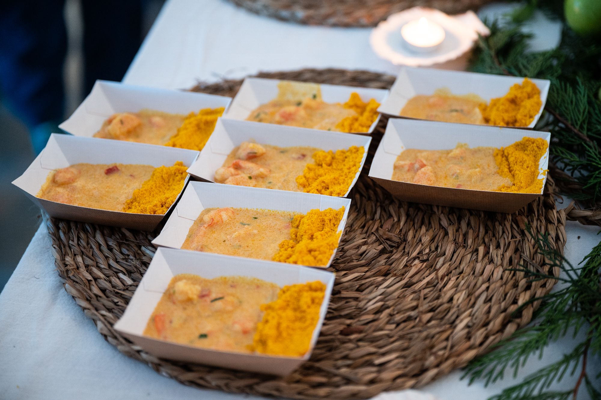 Seven trays of yellow seafood stew with a textured golden topping, arranged on a woven placemat, with soft candlelight in the background. - José Caldeira