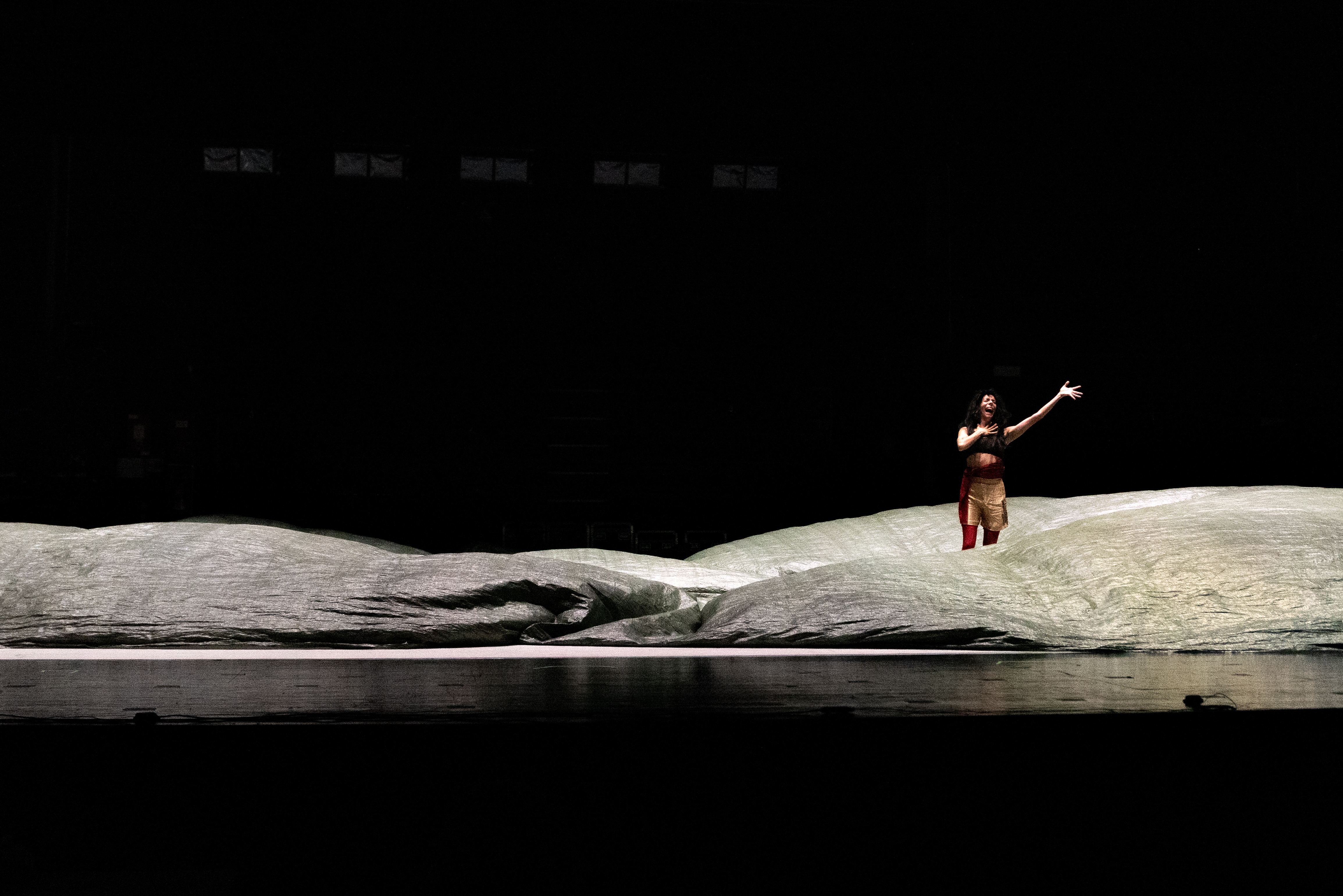 A lone performer stands on a dimly lit stage, surrounded by large, undulating fabric resembling waves. The scene conveys a sense of drama and solitude. - Walesca Timmen