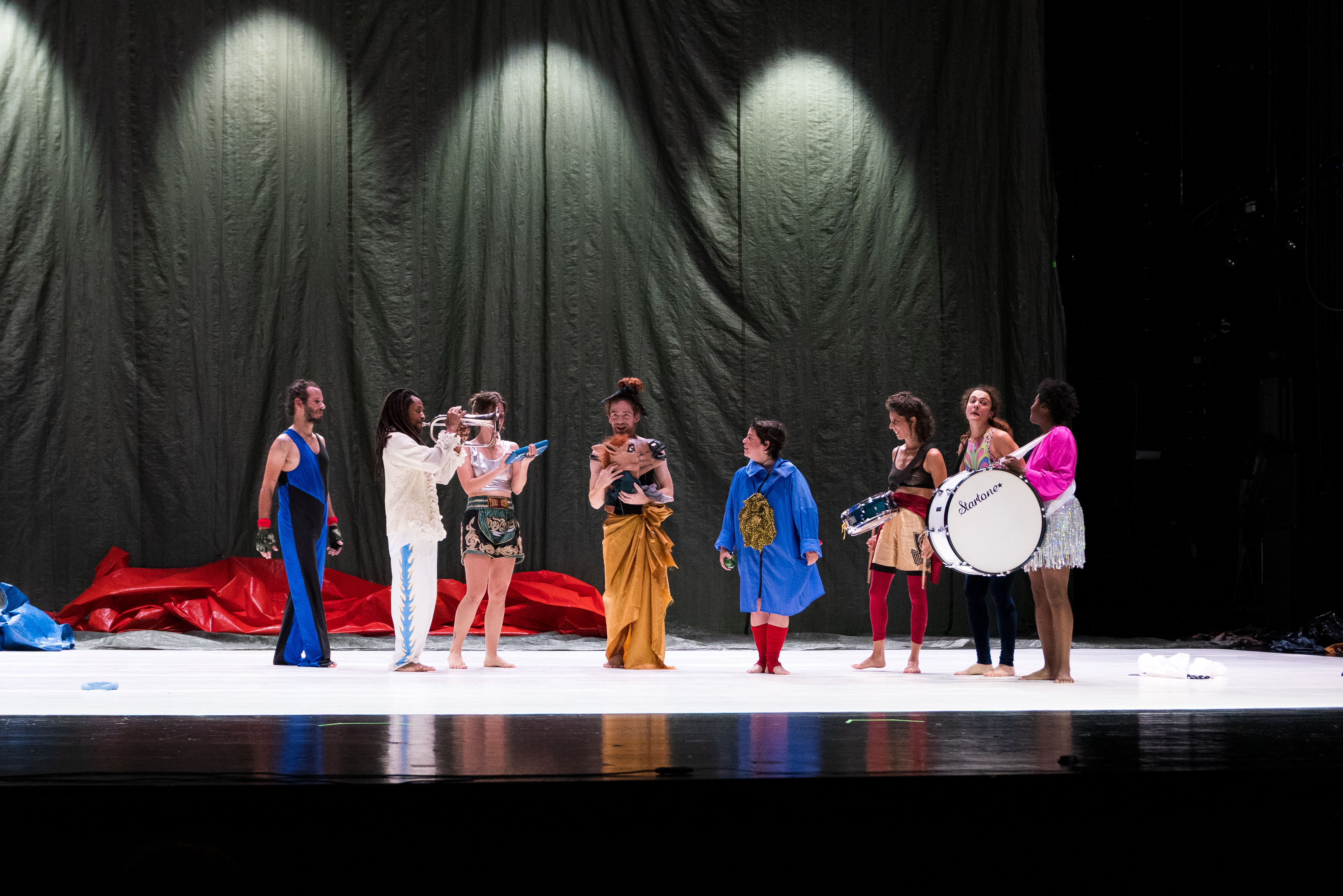 A group of eight people in colorful, eclectic costumes stand on a stage, some holding musical instruments, with a draped backdrop behind them. - Walesca Timmen