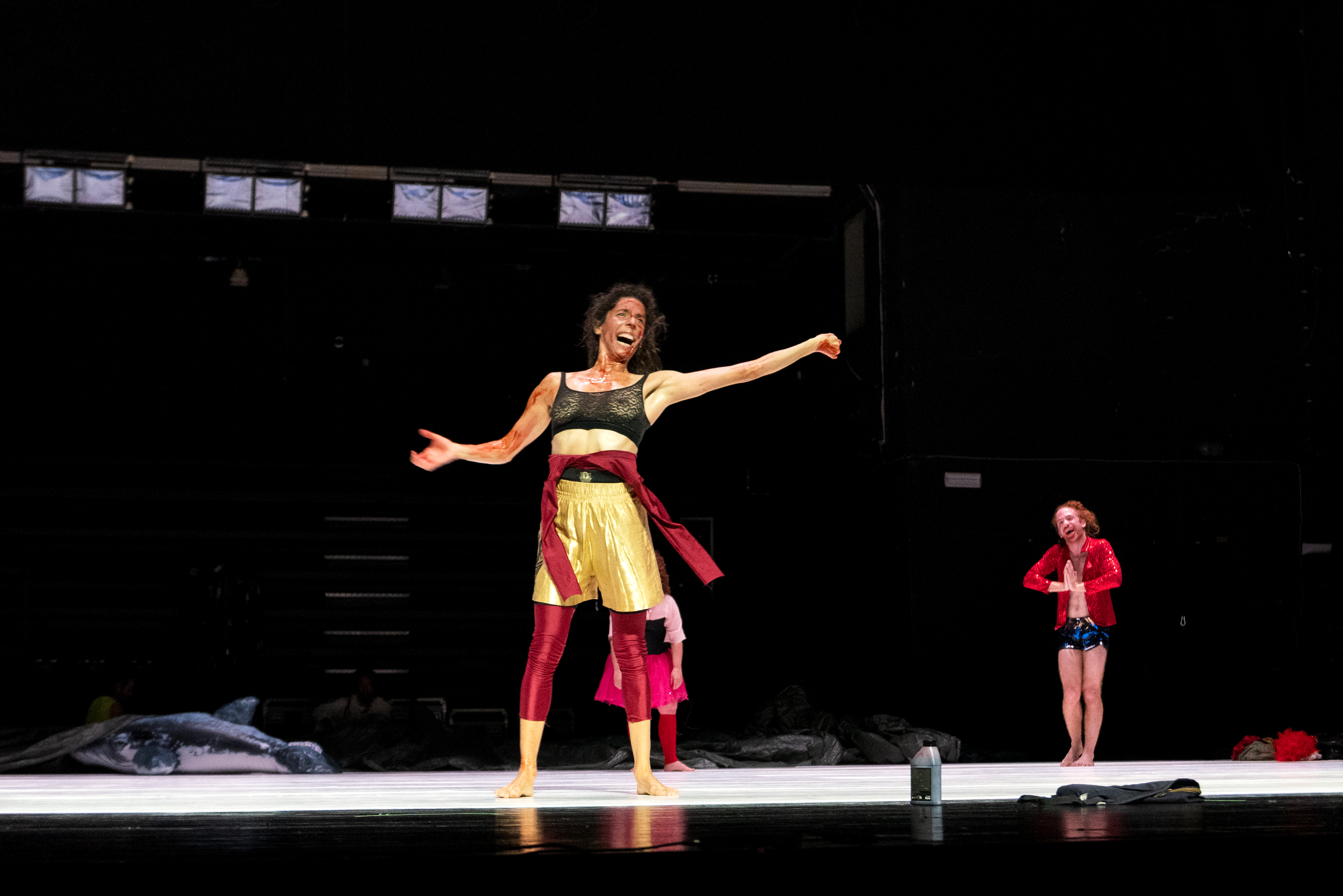 A performer on stage, center, wearing a glittery black top, gold skirt, and red sash, exudes energy and joy. Another dancer in a red jacket smiles in the background. - Walesca Timmen