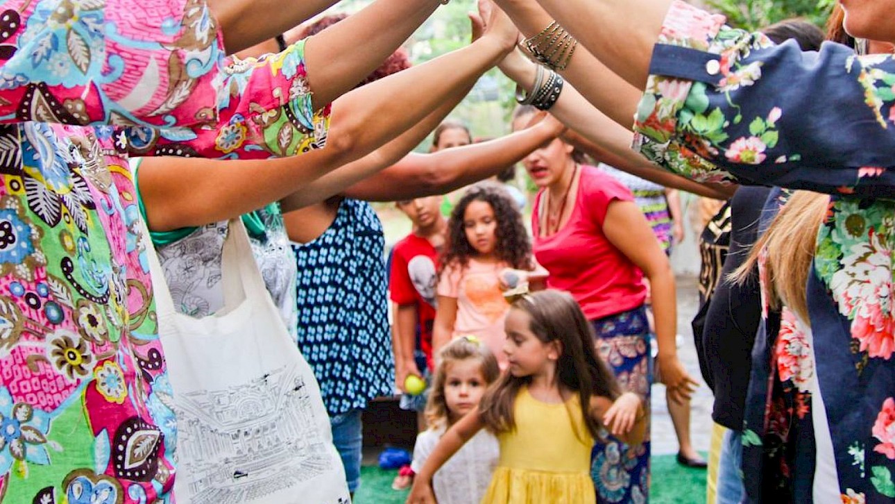 ALKANARA - Crianças passam por um arco formado pelos braços levantados de adultos num convívio ao ar livre. A cena é colorida e animada, transmitindo alegria e celebração. - ©lente materna