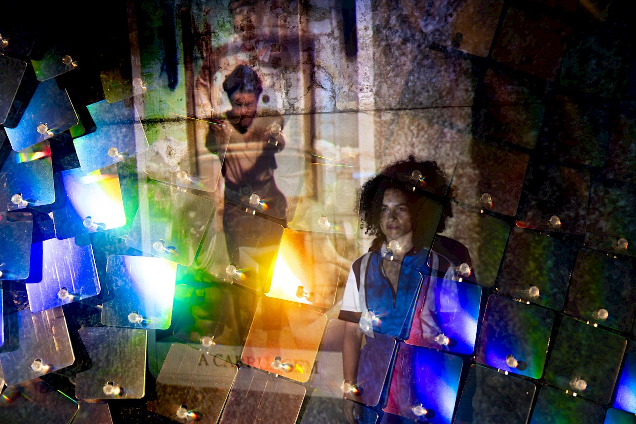 ALKANTARA - Colorful, layered collage with vibrant, overlapping lights and images. Two figures stand, overlaid by a grid of translucent squares, creating a dreamy aura. - ©Pedro Jafuno