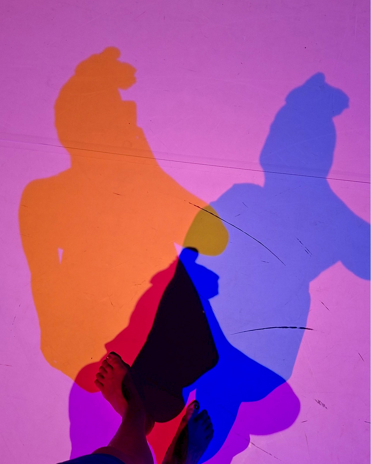 ALKANARA - Silhuetas coloridas projetadas no chão, misturando tons de laranja, azul e rosa. Pés descalços visíveis, sugerindo um ambiente divertido e artístico. - ©DR