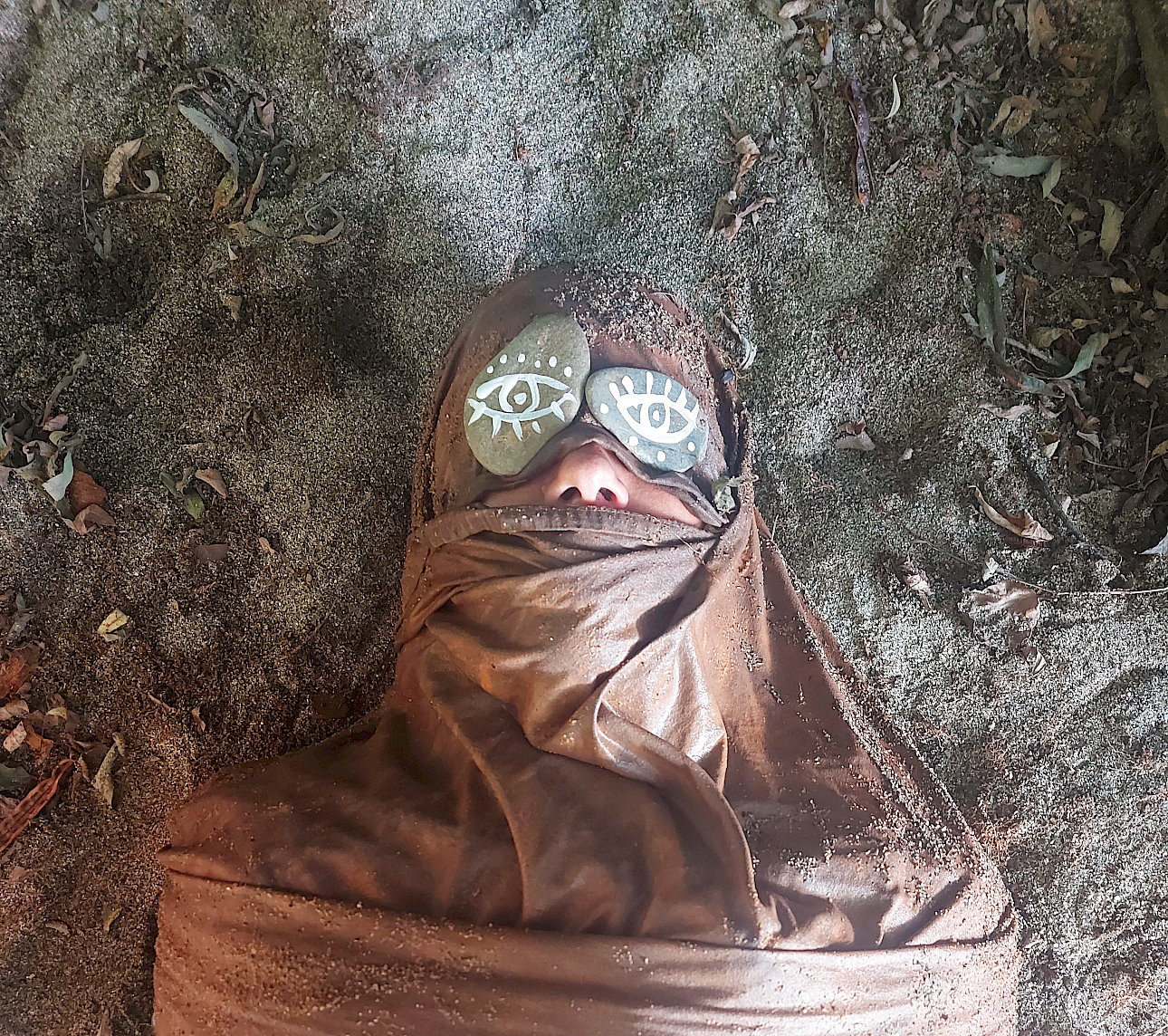ALKANARA - A person is partially buried in dirt, wrapped in brown fabric, with painted stones depicting eyes placed over their closed eyes. The setting is earthy and serene. - ©AND Lab