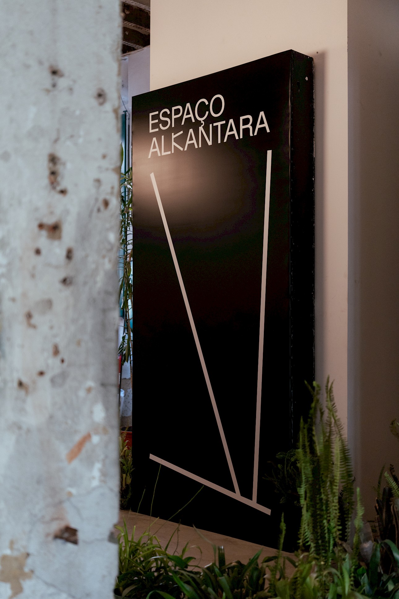 ALKANTARA - Black sign with white text reading "ESPAÇO ALKANTARA" against a textured wall, surrounded by green plants, conveying a modern, artistic vibe. - ©Joana Linda