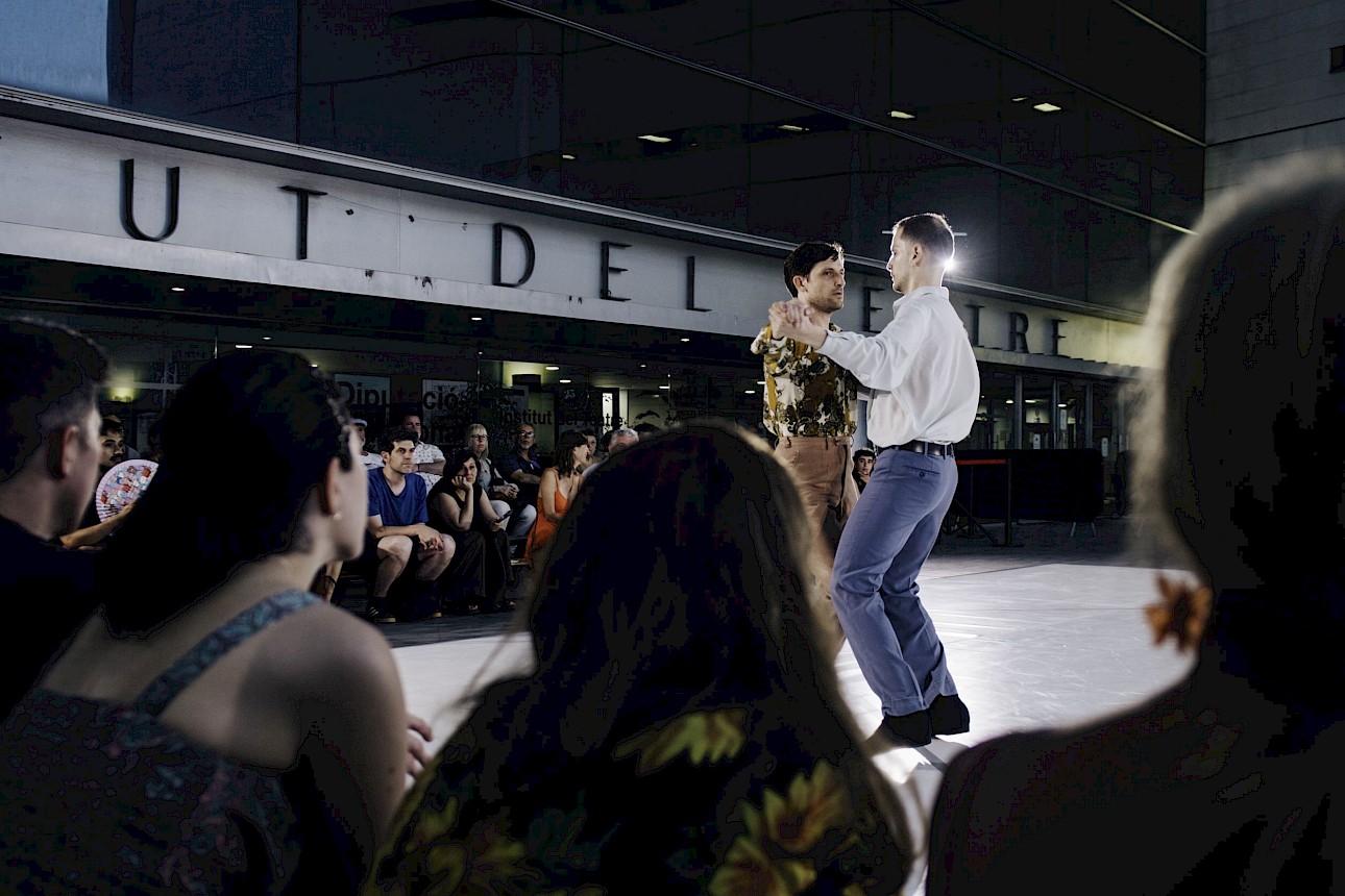 ALKANTARA - Two men partner dancing on an outdoor stage at night, captured from behind a blurred audience. A modern glass building with the text "Institut del Teatre" is visible in the background. - ©Alice Brazzit