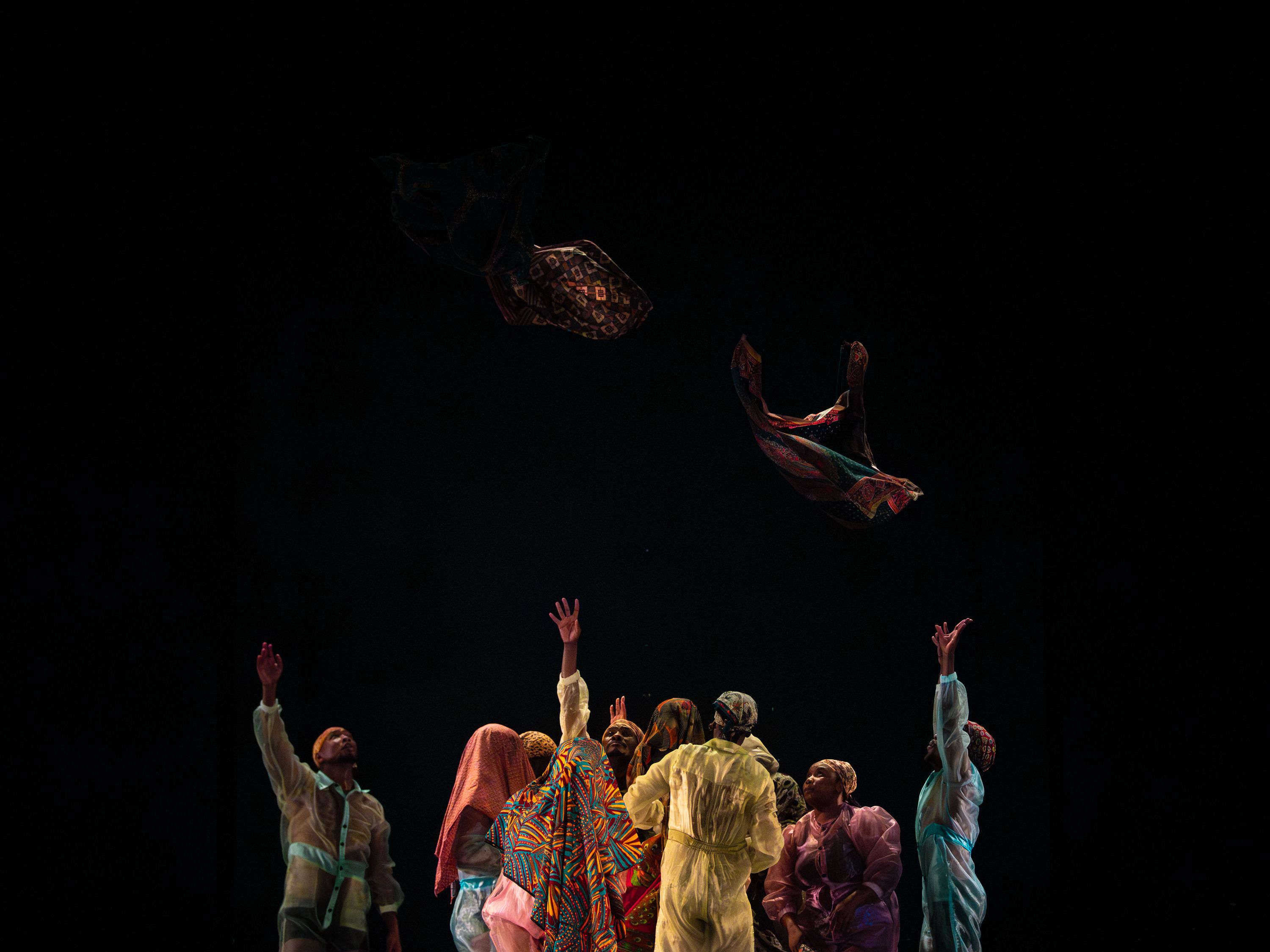 A group of people wearing colorful clothing stand in a circle, reaching upward as patterned fabric pieces float above them against a dark background. - Andreas Hagenbach