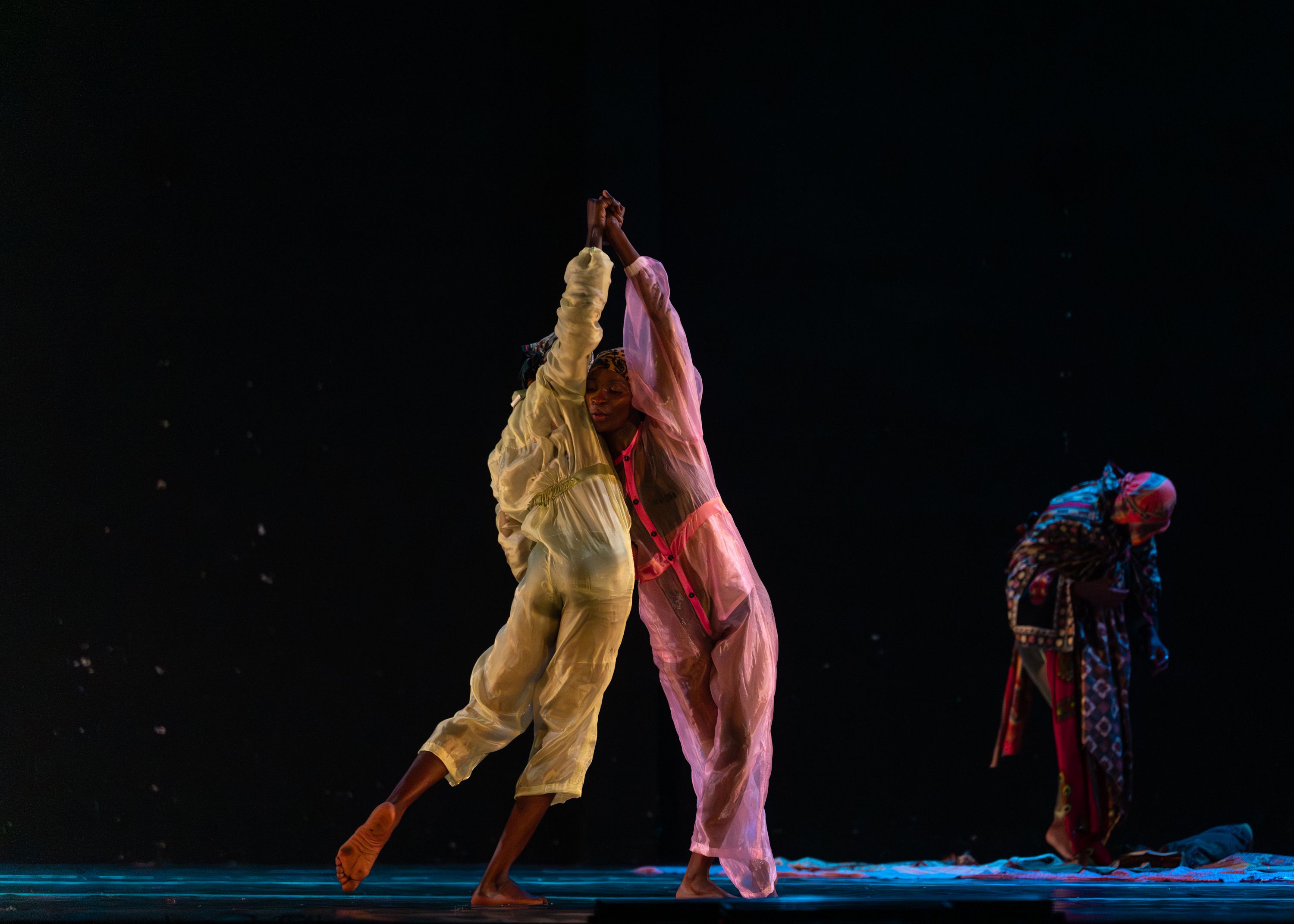 Three dancers in patterned clothing and headwraps against a dark background. Two of them are embraced and balanced with linked, high hands. - Andreas Hagenbach