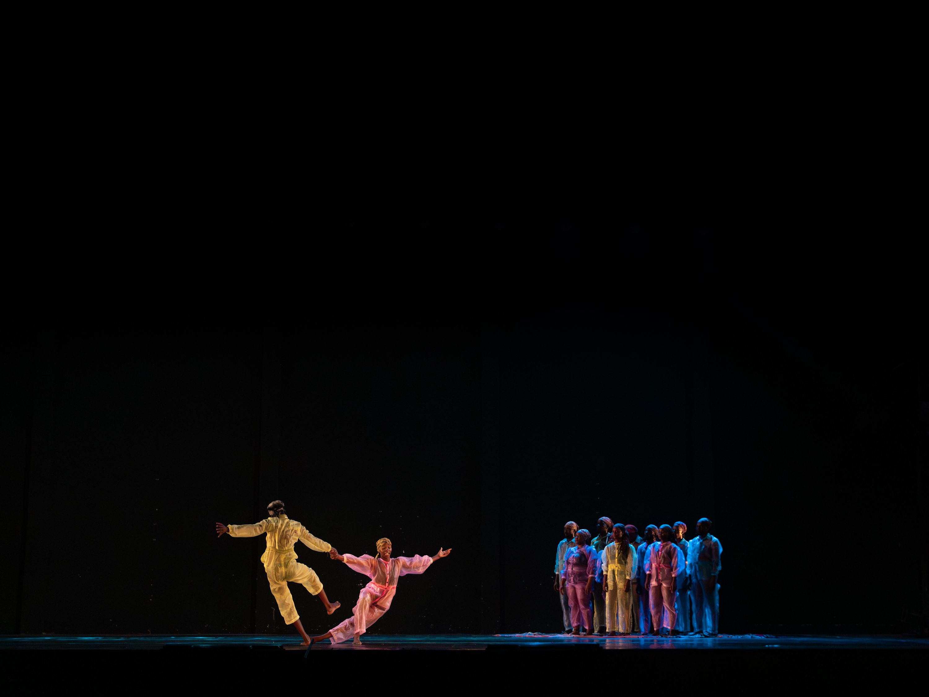 Two dancers in yellow and pink clothing hold hands and lean away from each other in a dynamic pose, while a larger group watches from the side against a vast dark background. - Andreas Hagenbach