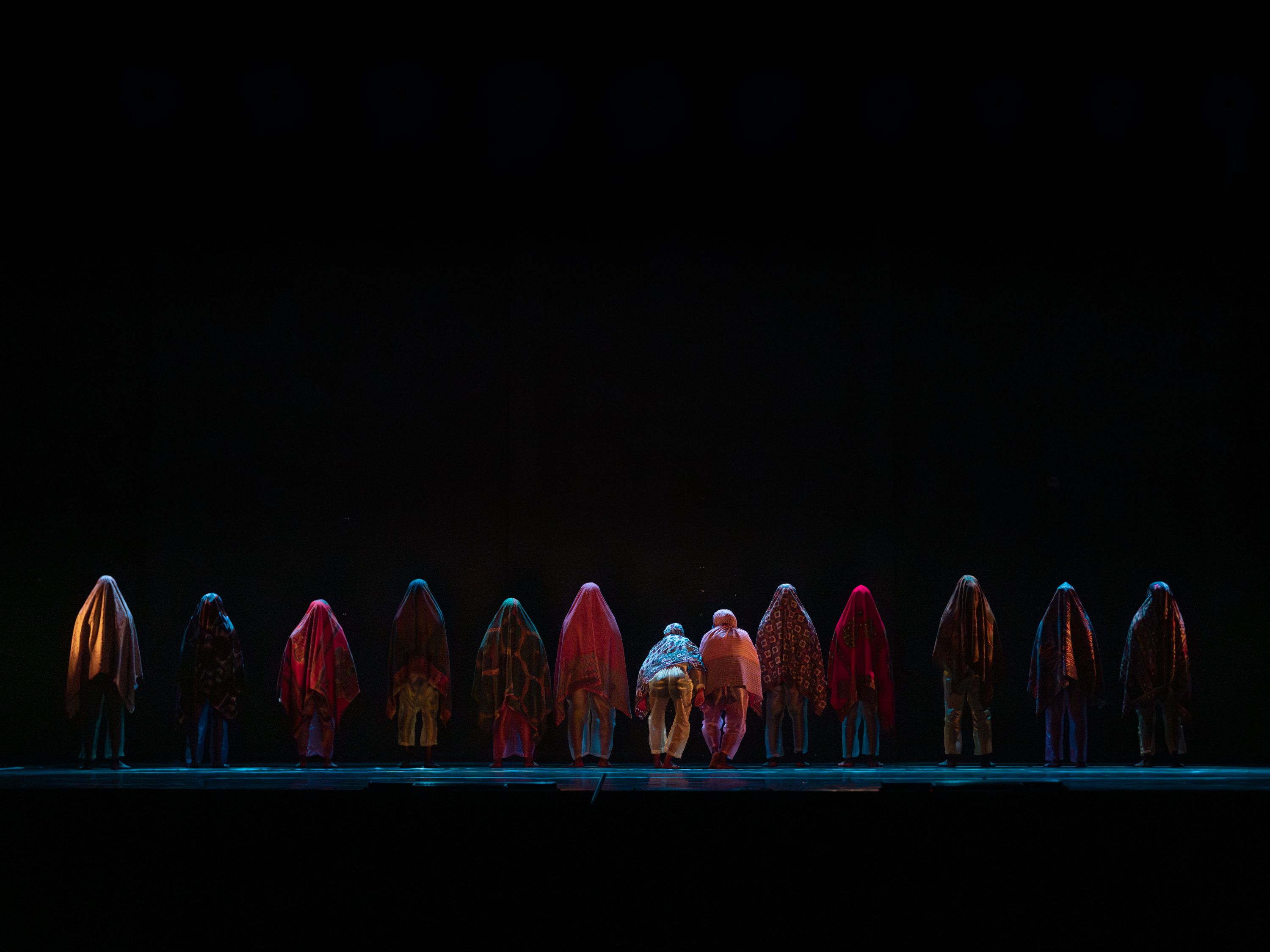 A line of people covered in colourful, patterned fabrics stand across a stage against a vast dark background. - Andreas Hagenbach