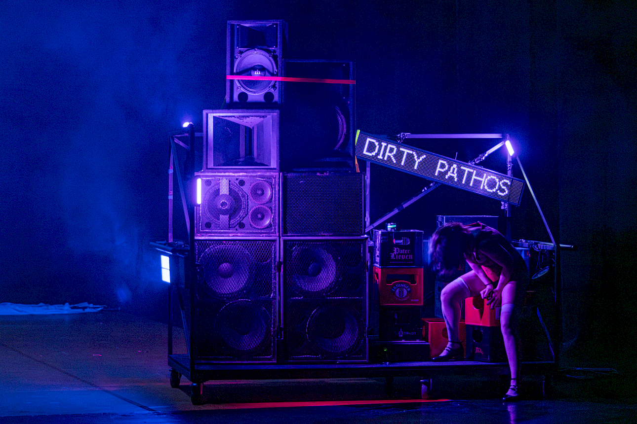 ALKANTARA - A performer sits on a mobile platform next to a large stack of loudspeakers under blue and purple stage lighting. An electronic LED sign displays the words "DIRTY PATHOS". - ©Mayra Azzi