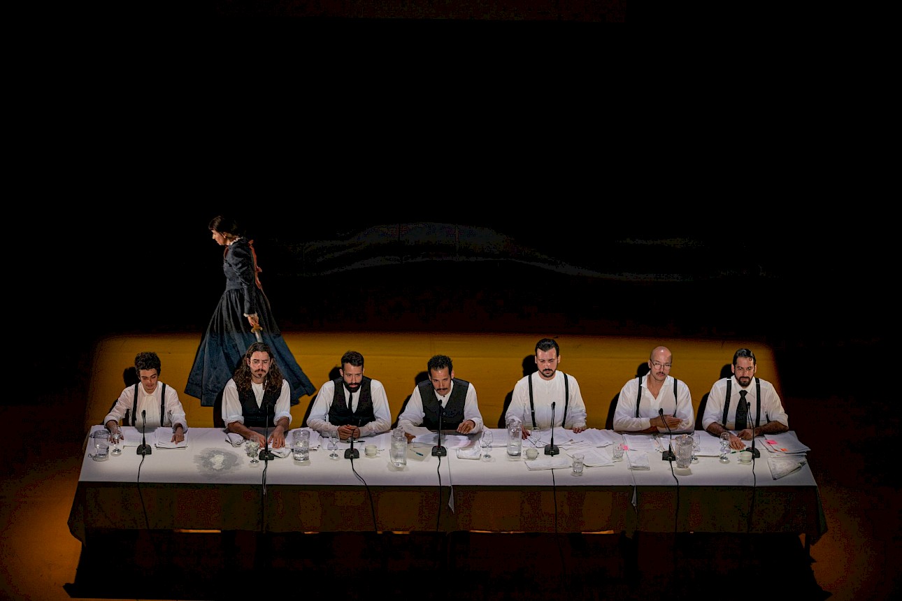 ALKANTARA - Seven men wearing white shirts with dark waistcoats or braces are seated in a row behind a long table on an illuminated stage. Microphones and papers are set out in front of each person. In the dimly lit background, a figure in a long dark dress walks in profile. - ©Mayra Azzi