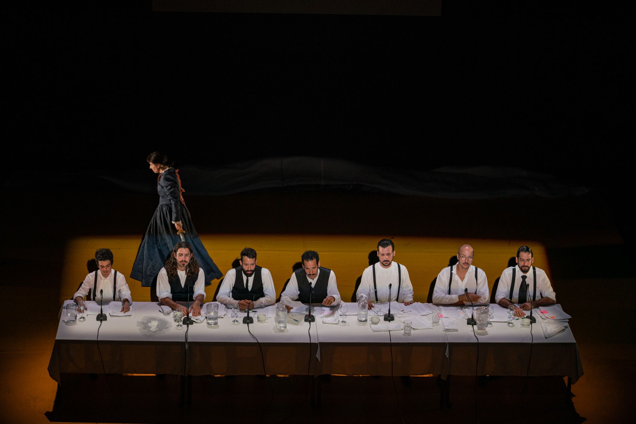 Seven men wearing white shirts with dark waistcoats or braces are seated in a row behind a long table on an illuminated stage. Microphones and papers are set out in front of each person. In the dimly lit background, a figure in a long dark dress walks in profile. - Mayra Azzi