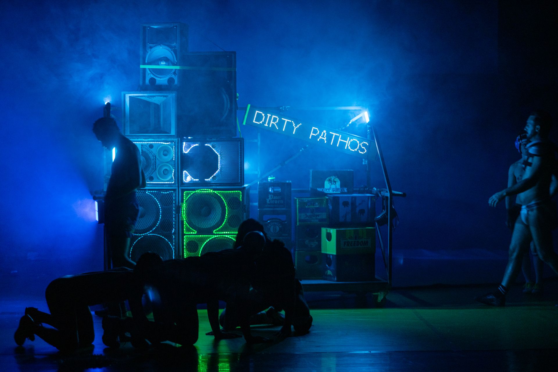 On a smoky stage with blue lighting, several silhouetted figures crawl on the floor in front of a stack of loudspeakers and crates. A light-up sign displays the words 'DIRTY PATHOS'. - Mayra Azzi