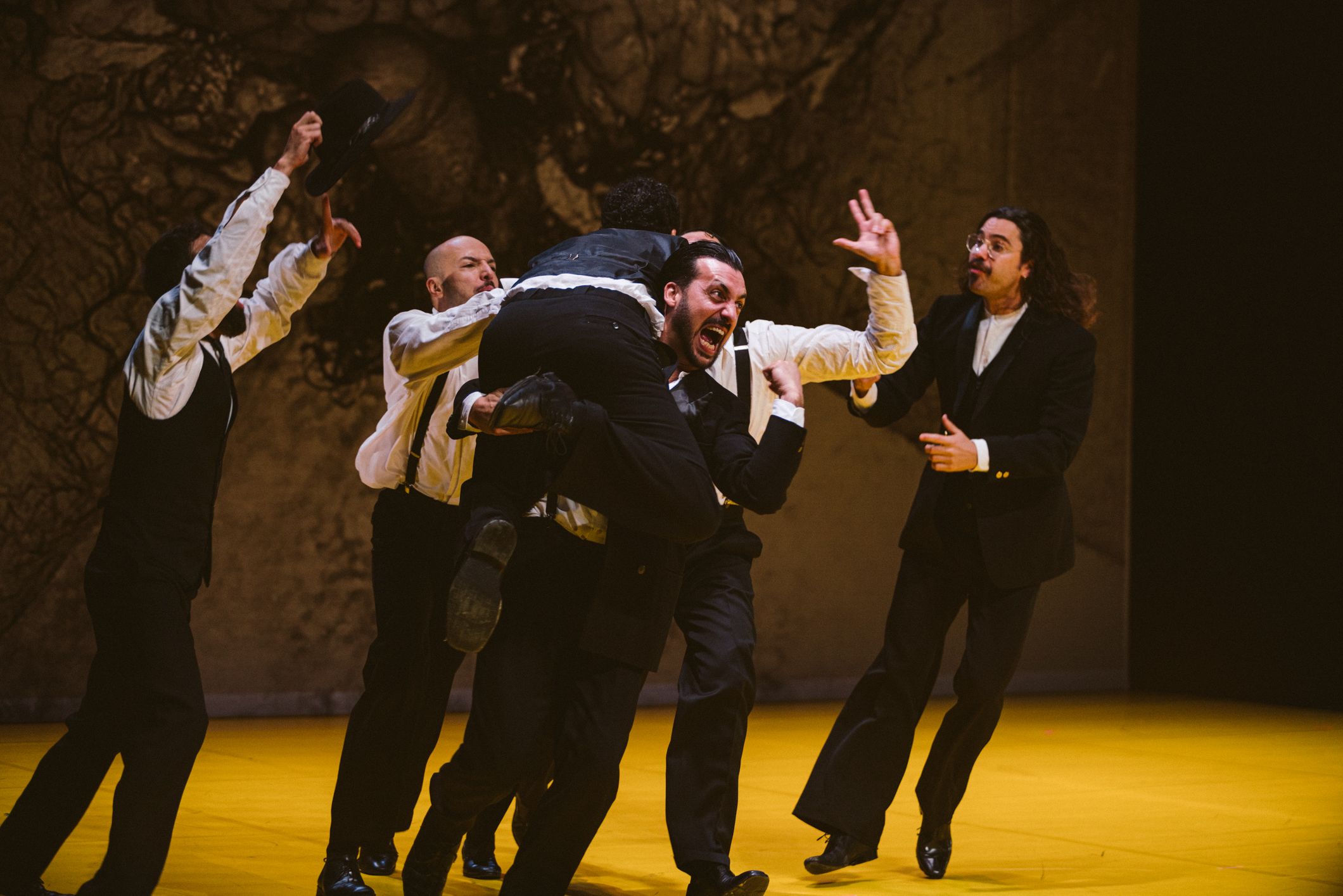 Vários atores vestidos com camisas brancas e calças pretas numa cena de teatro física e intensa sobre um palco amarelo. Um deles é carregado nos braços do grupo enquanto outro grita de forma expressiva. - Mayra Azzi
