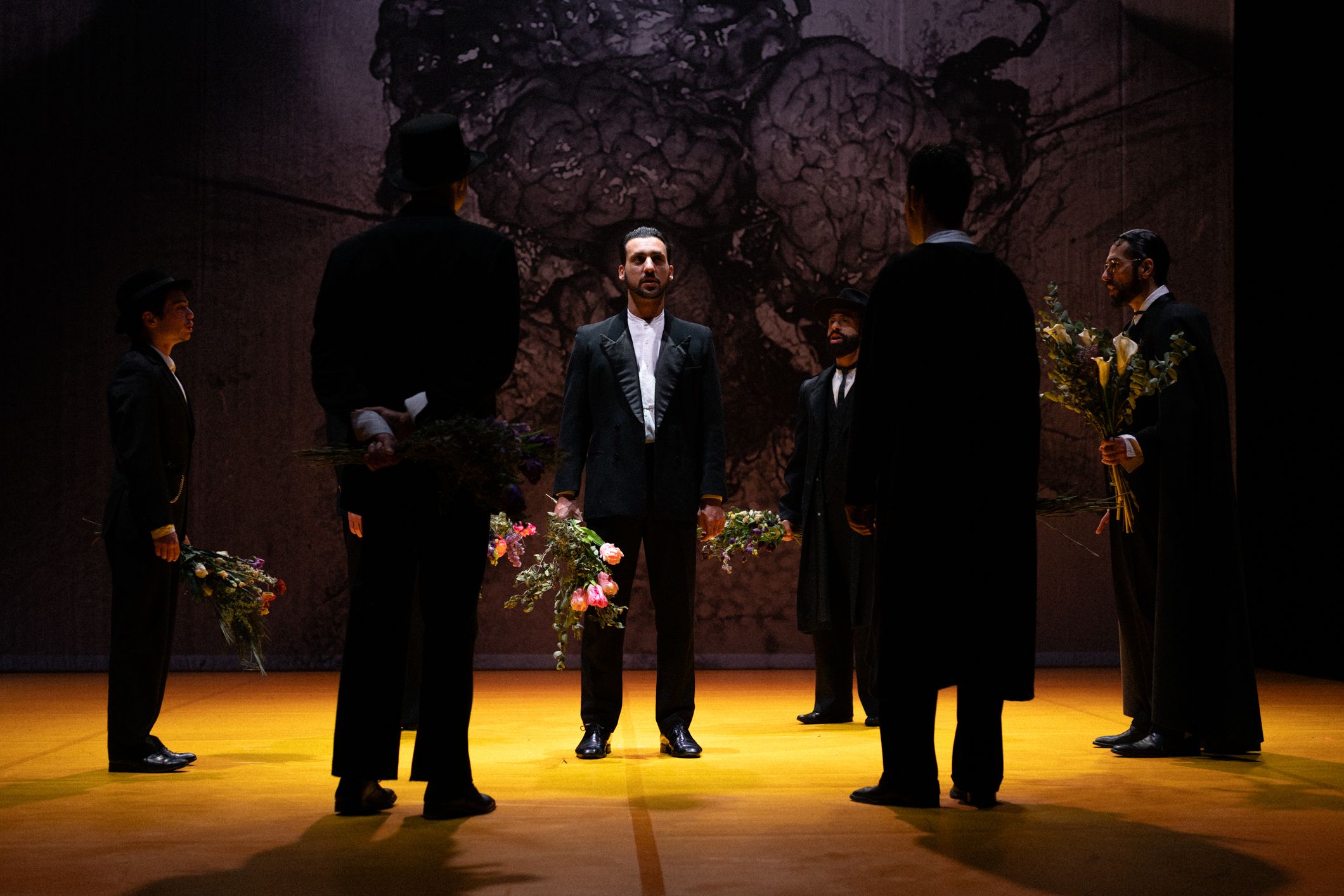 A group of men dressed in dark suits stand in a circle on a brightly lit yellow stage. The man in the centre faces forward, while the others hold bouquets of flowers. In the background, a large textured backdrop displays a dark abstract image. - Mayra Azzi