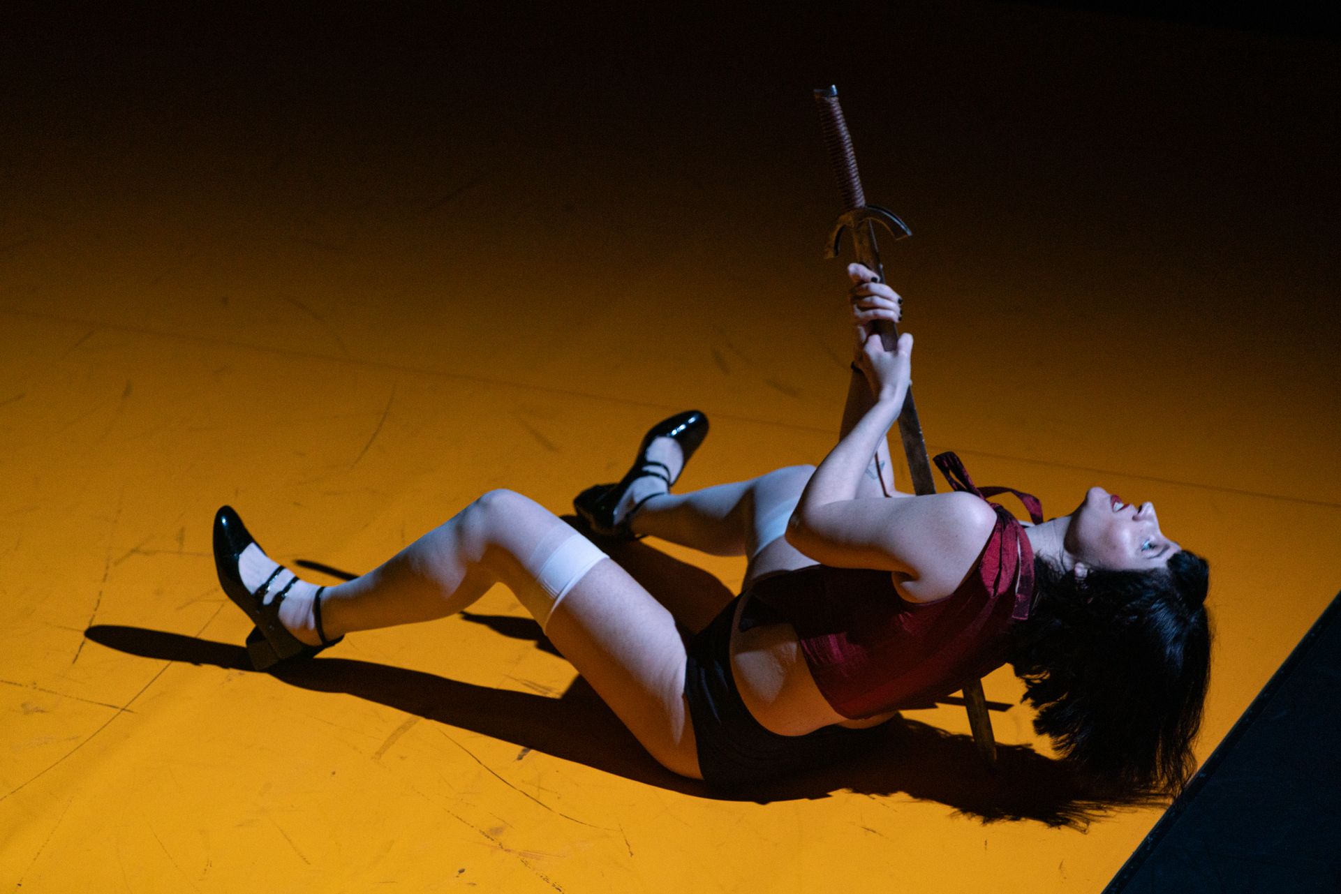 A short-haired person lies on their back on a brightly lit yellow floor, holding a sword vertically against their chest. They are wearing a burgundy corset, black shorts, high white stockings, and black heeled shoes. - Mayra Azzi