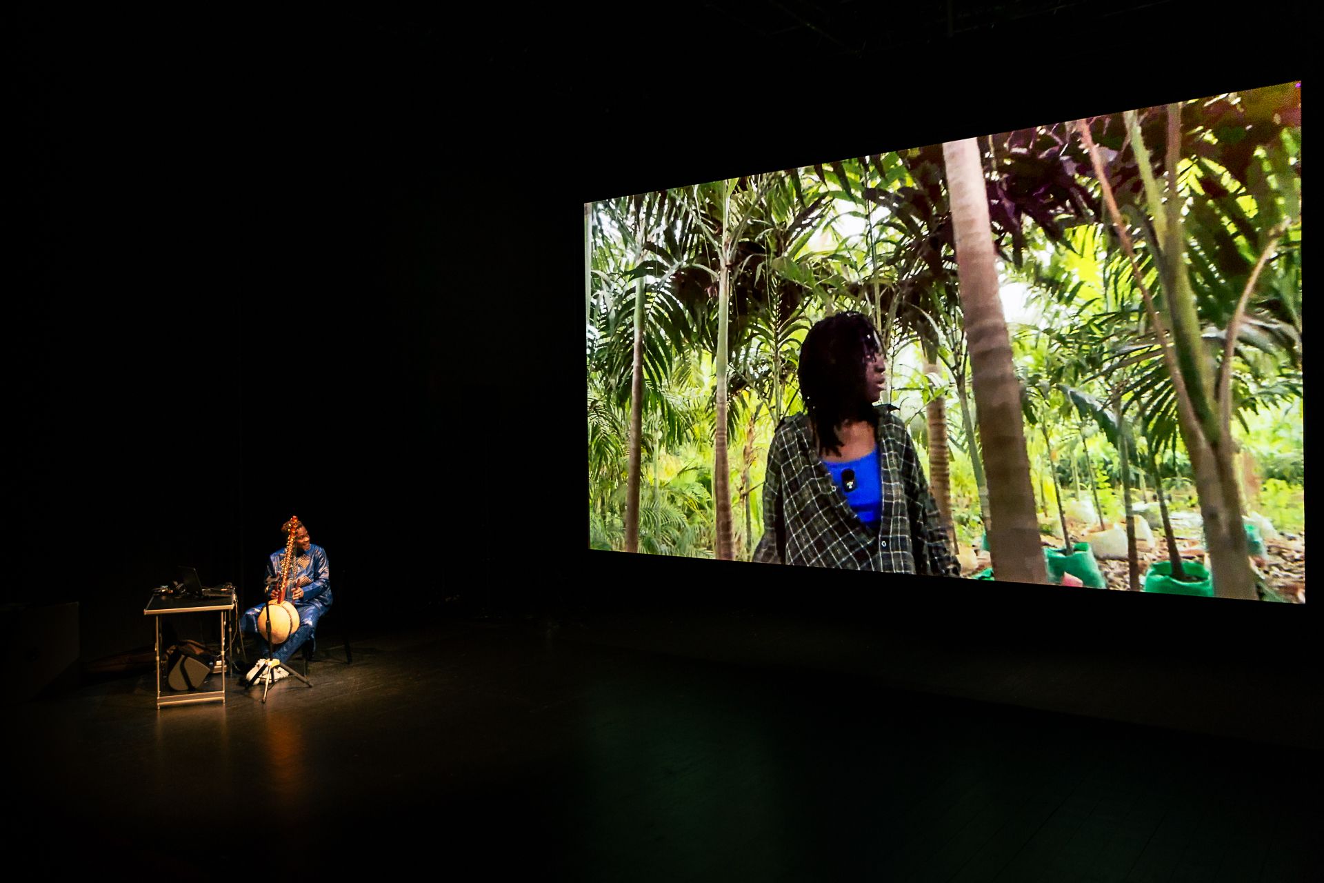 A wide shot of a dark stage with a musician playing a kora on the left. In the background, a large screen displays a woman in a plaid shirt among palm trees. - Claudia Ndebele