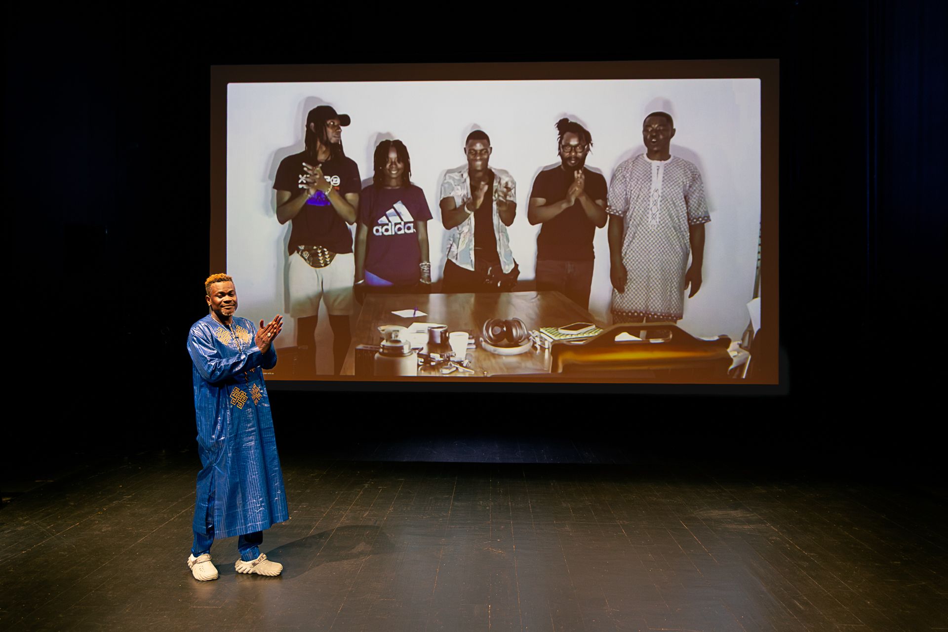 A man in a blue tunic claps on stage in front of a large projection screen displaying five people clapping. - Claudia Ndebele