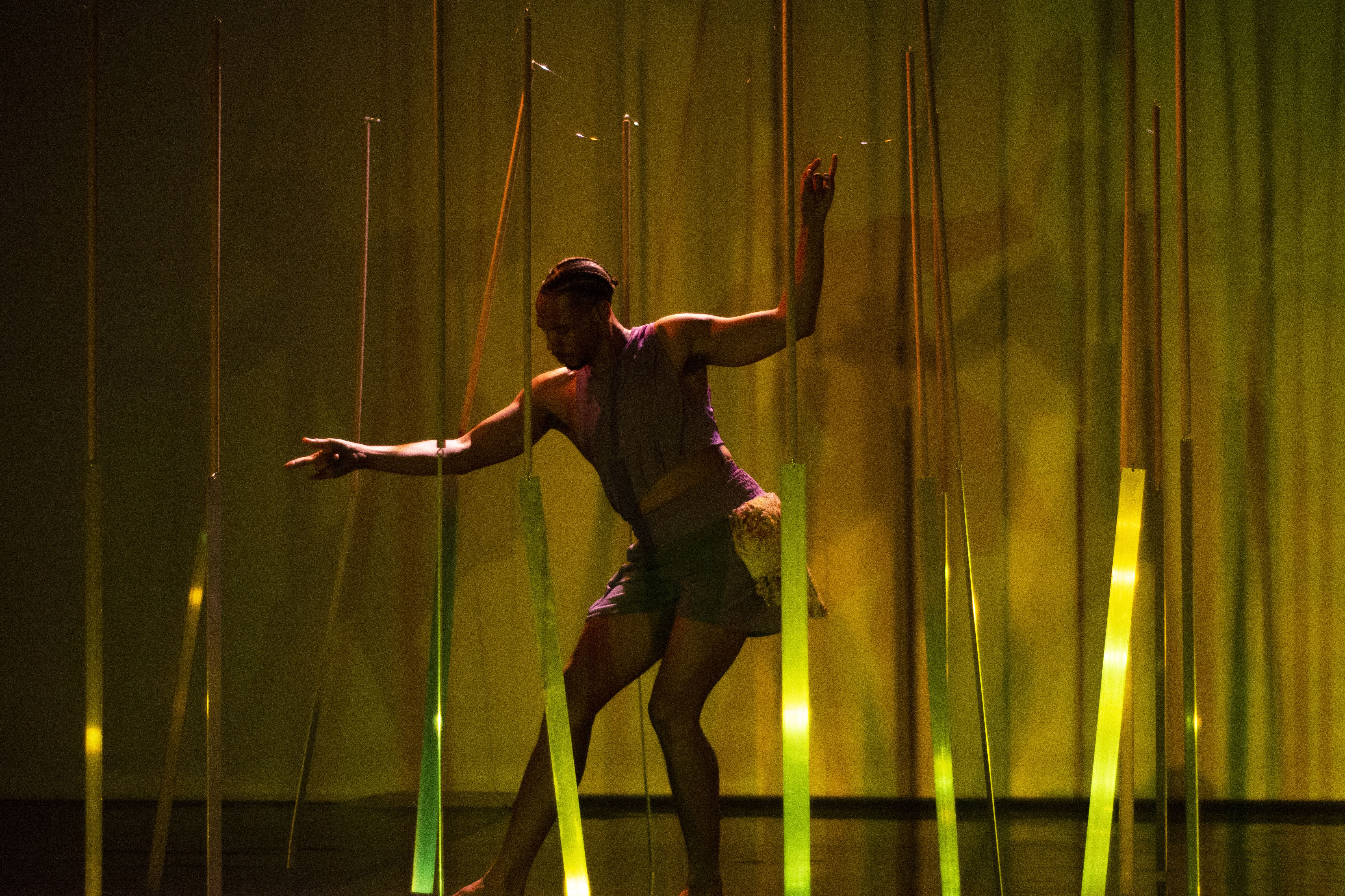 Uma pessoa com tranças no cabelo dança num palco, flectindo os joelhos e estendendo os braços. Move-se por entre várias tiras verticais que brilham com uma luz verde e amarela, sob um fundo quente. - Rui Palma