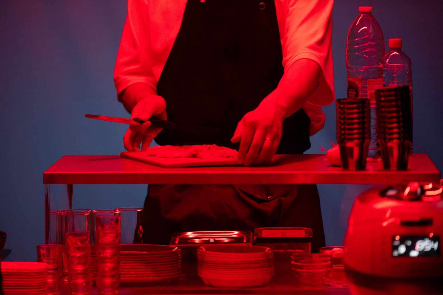 A person wearing a white t-shirt and black apron holds a knife and cuts food on a chopping board at a counter. The entire scene is lit by a strong red light against a blue background. - Bea Borgers