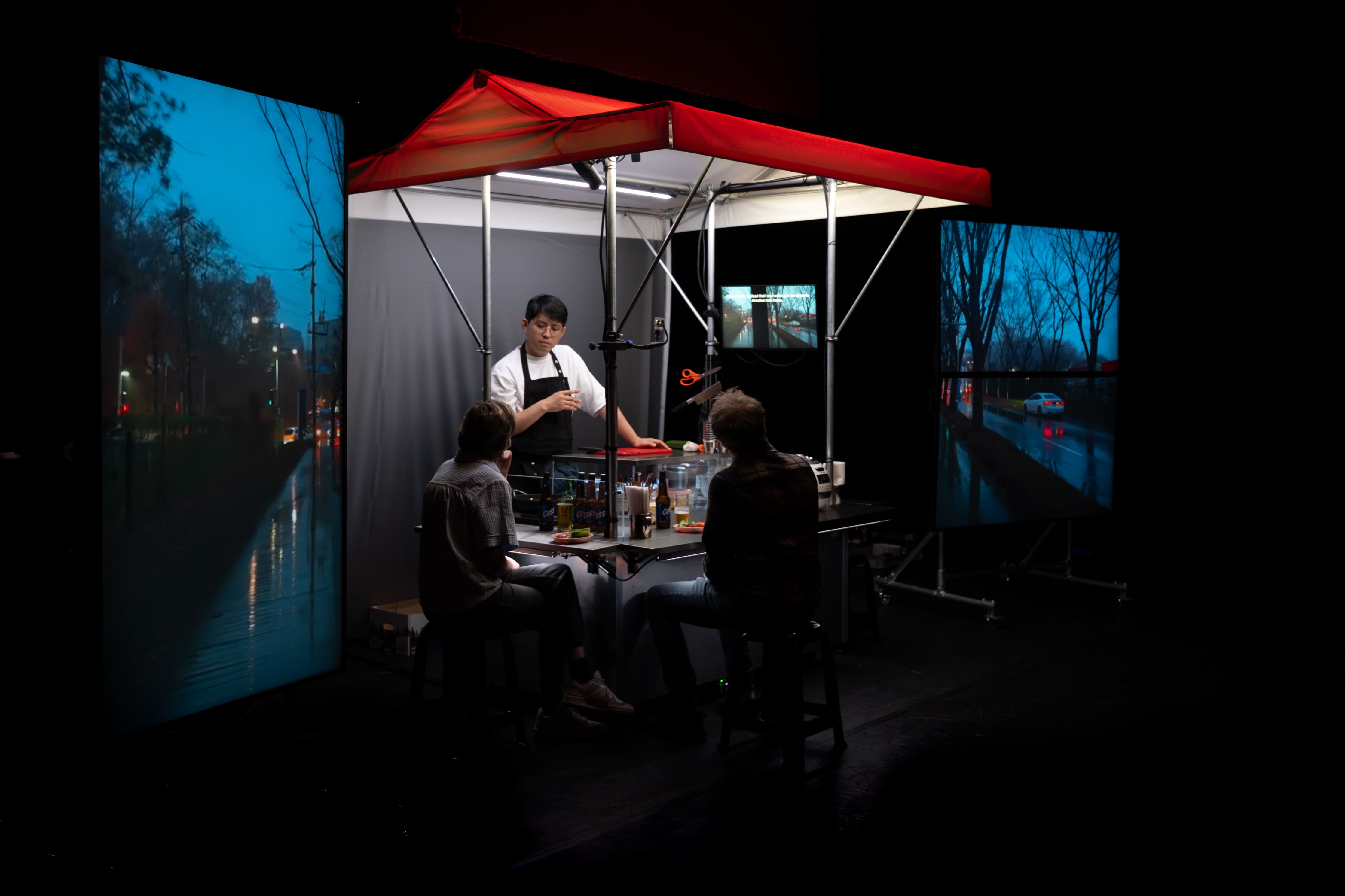 A closer view of a stage art installation, focused on a Korean food stall under a red awning. A chef in a black apron talks to two customers seated at the counter. The stall is framed by two large vertical LED screens displaying rainy, night-time cityscapes, reflecting the city lights. - Bea Borgers