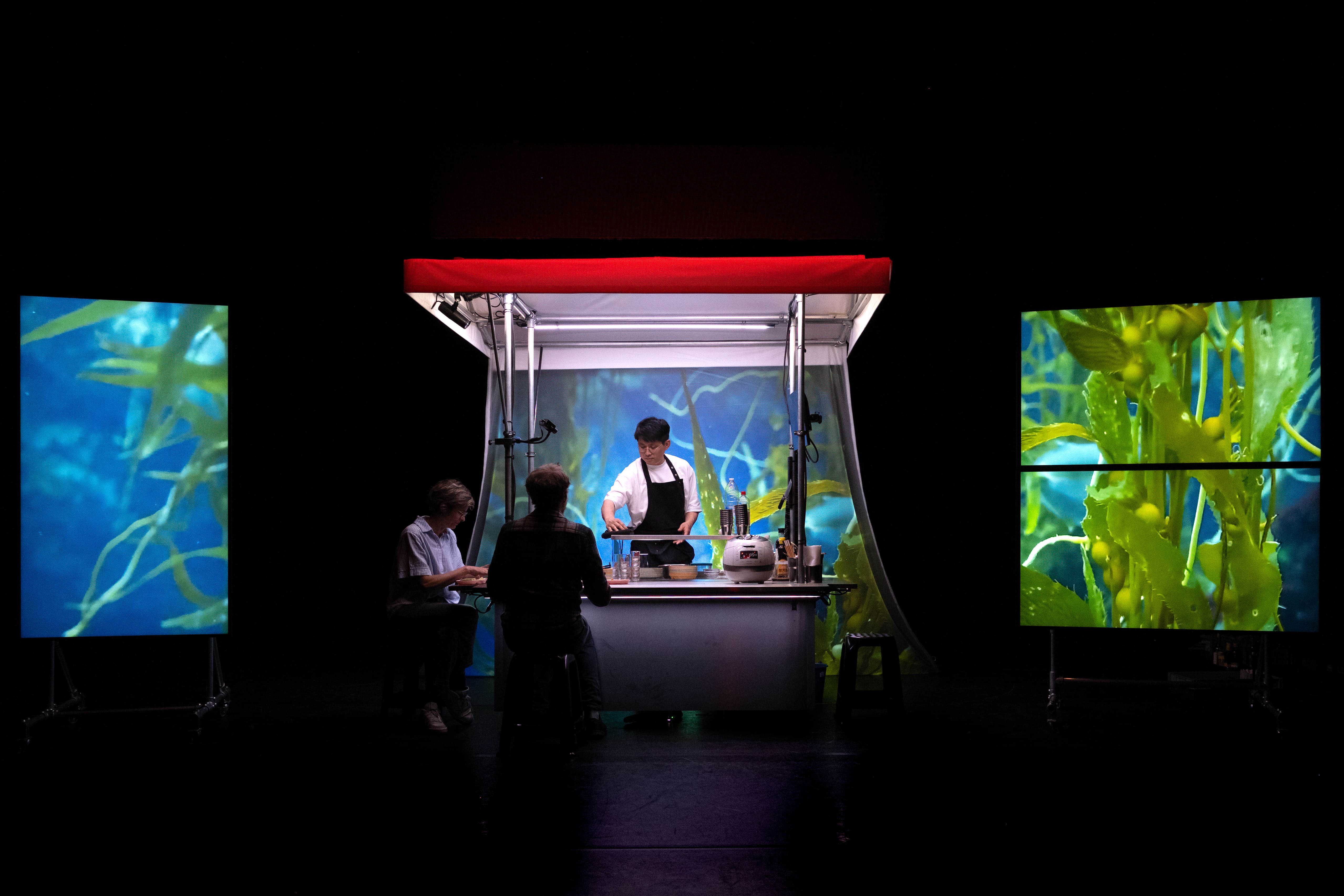A stage art installation, showing a lit Korean food stall with a chef and two seated customers. The background of the stall and two large side LED screens display vibrant underwater images of green and blue kelp, contrasting with the dark stage and the stall's red awning. - Bea Borgers