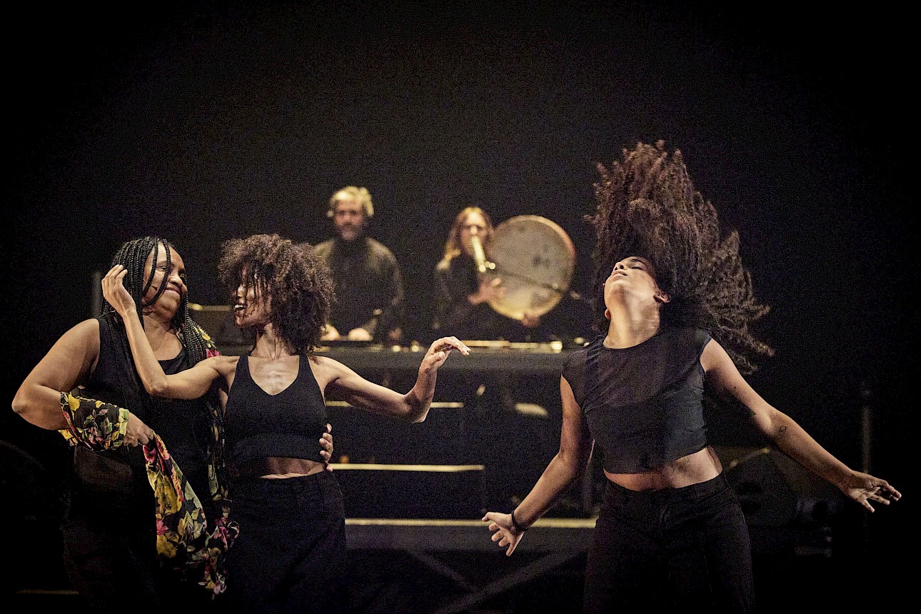 ALKANTARA - Três mulheres dançam com energia em primeiro plano, enquanto dois músicos atuam no palco, ao fundo. - ©Christophe Raynaud De Lage