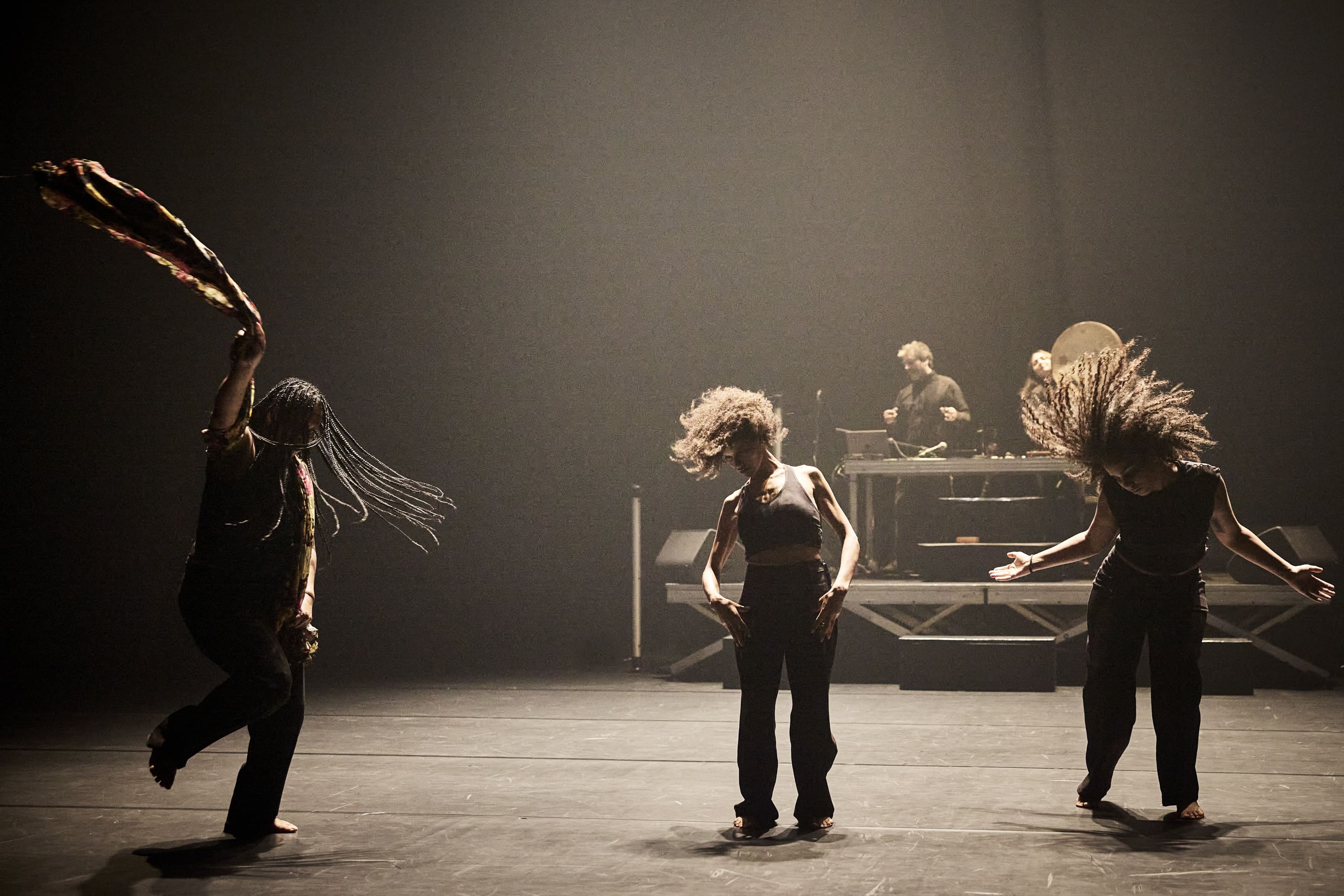 © Christophe Raynaud De Lage Three people dance energetically on a dimly lit stage, with a DJ performing on a raised platform in the background. - Christophe Raynaud De Lage