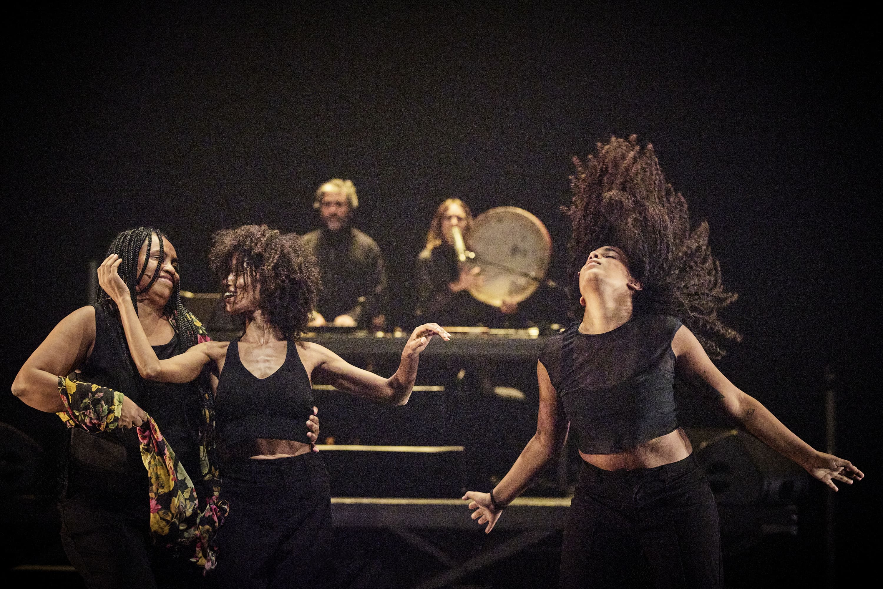 © Christophe Raynaud De Lage Three women dance energetically in the foreground while two musicians perform on stage in the background. - Christophe Raynaud De Lage