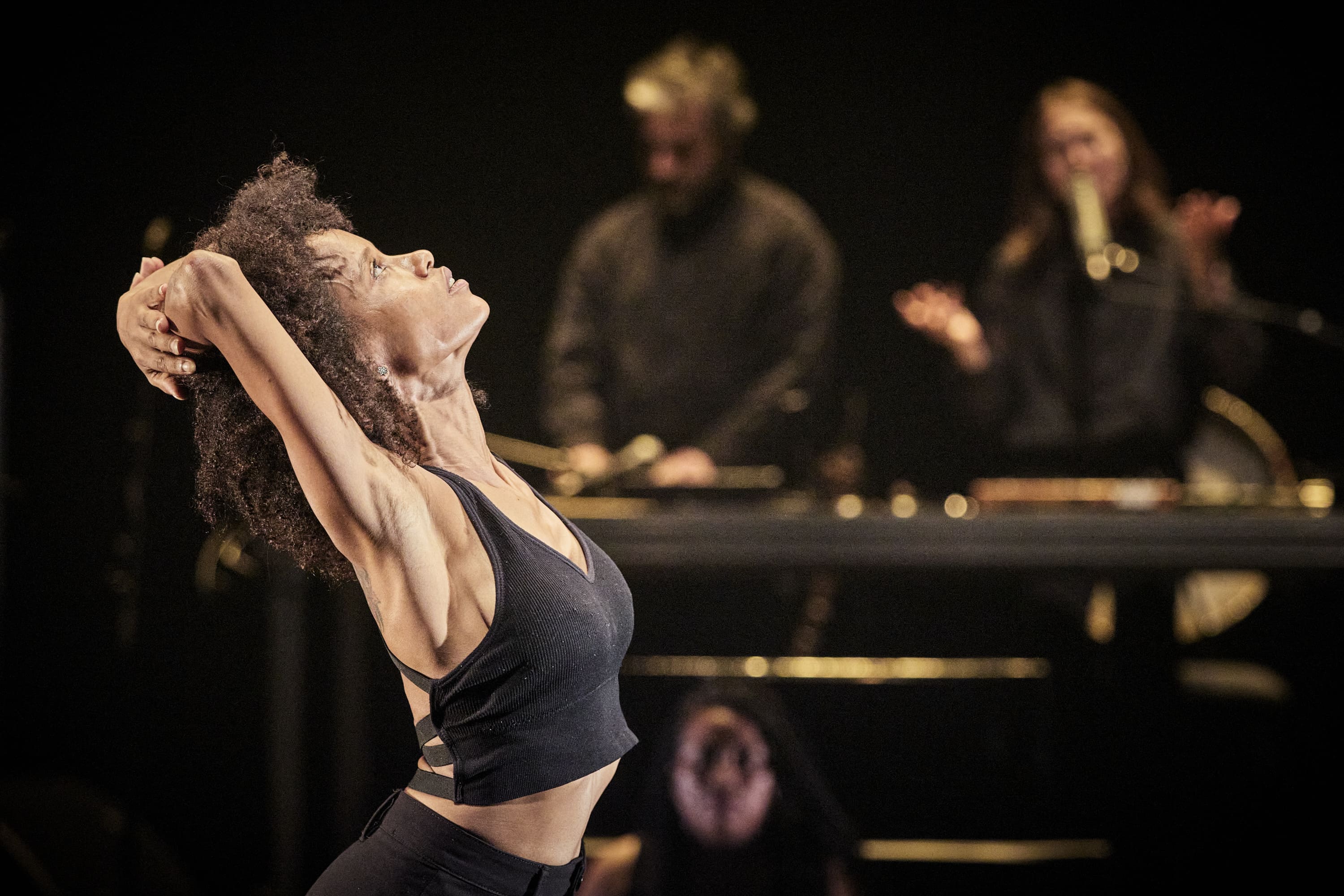 © Christophe Raynaud De Lage A Black woman with an afro hairstyle, wearing a black crop top, bends her torso backwards and looks upwards, her hands interlaced behind her neck in an expression of release or strain. In the background, out of focus, are two musicians with percussion instruments and keyboards, on a stage with soft lighting. - Christophe Raynaud De Lage