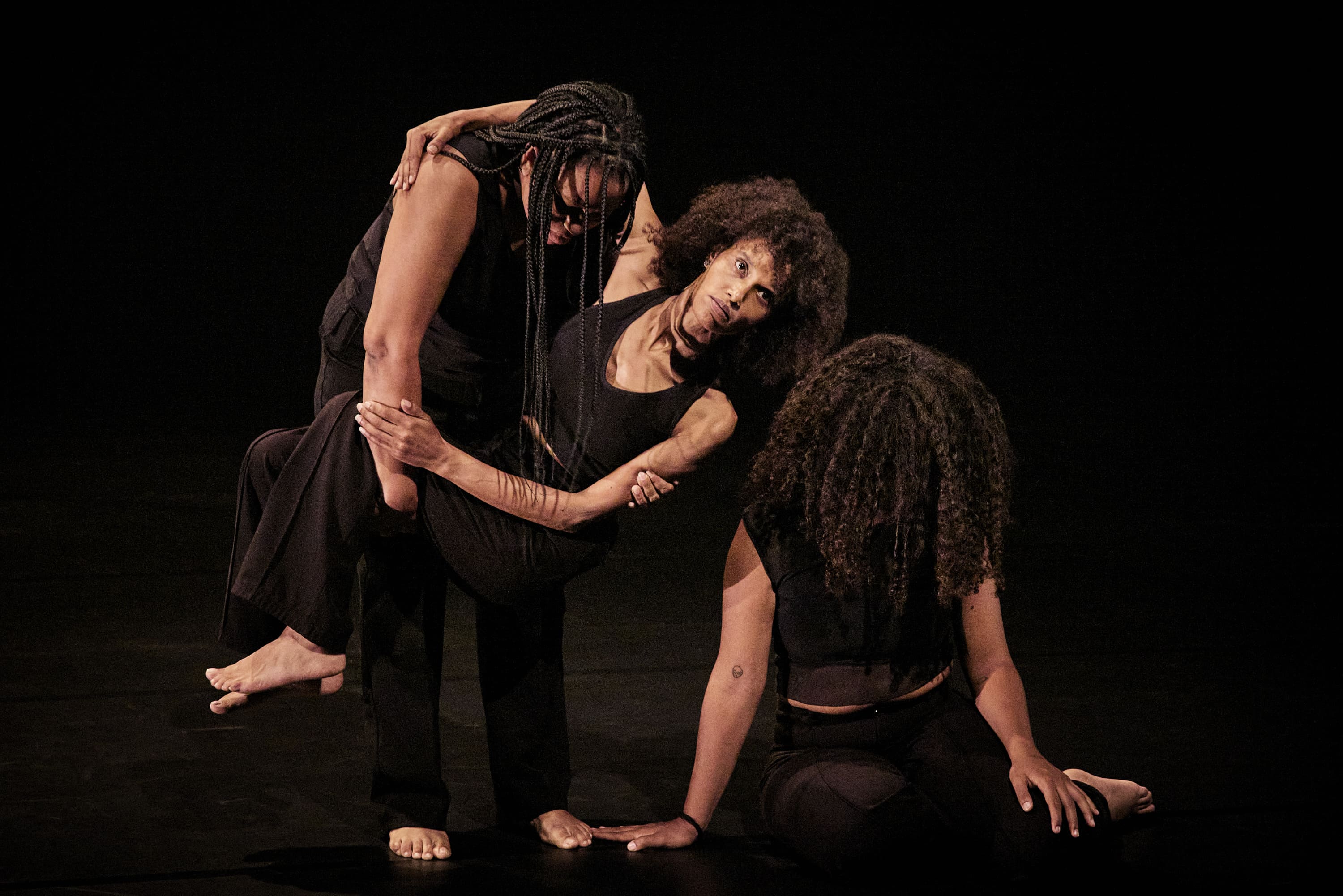 © Christophe Raynaud De Lage Three Black women in black clothes on a dark stage. In the centre, a woman with an afro hairstyle is being supported and lifted by another woman with braids. The third woman, with curly hair, sits on the floor in the foreground, her face hidden by her hair, propping herself up on her hands. The composition suggests mutual support and helplessness. - Christophe Raynaud De Lage