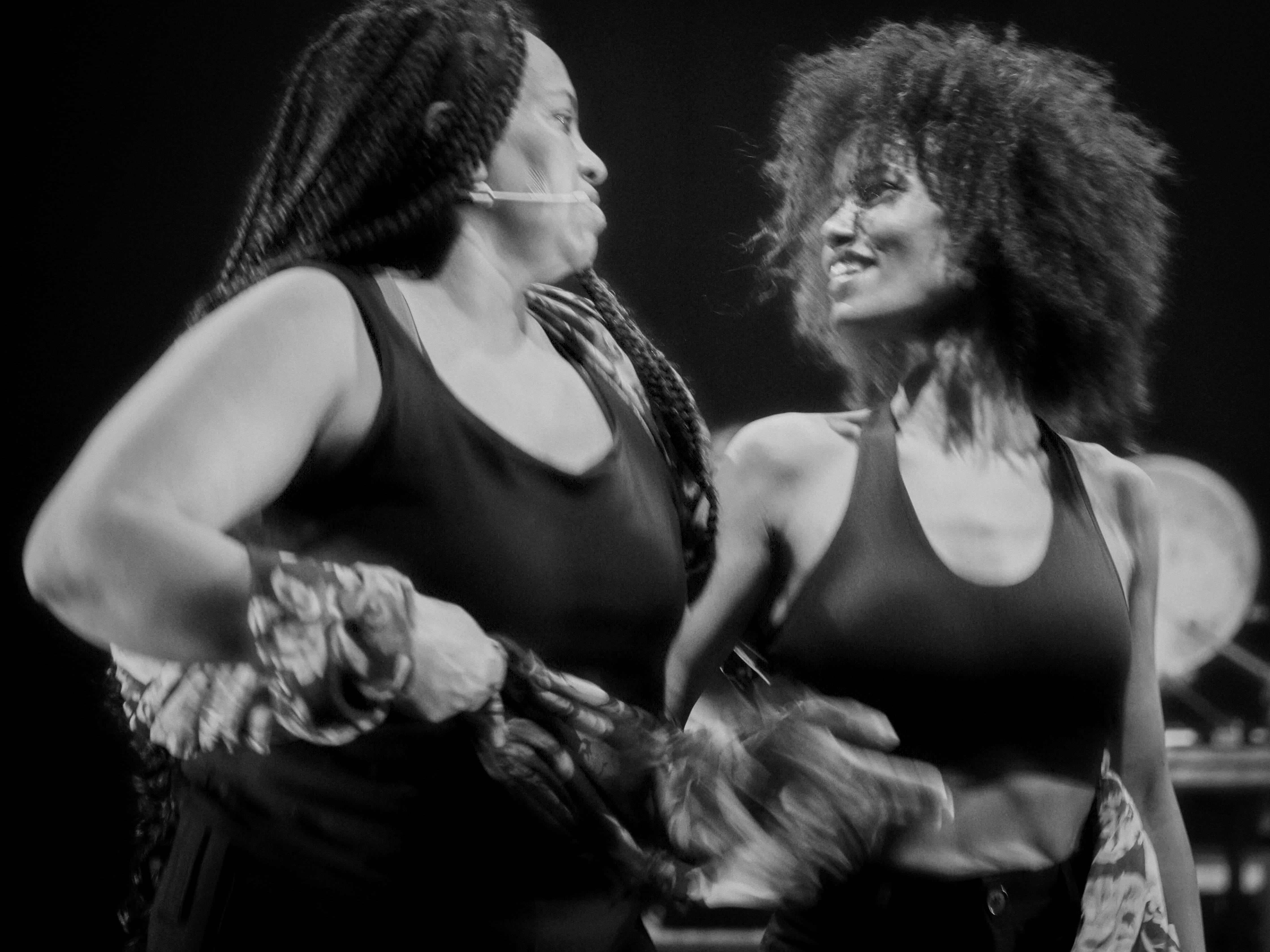 © Lea Skayem A medium black and white photograph shows two Black women on a stage, looking intensely and smiling at each other. Both are wearing black crop tops. The woman on the left has braids and a headset microphone, and holds a patterned fabric in her hands. The woman on the right has an afro hairstyle and wears a strappy top. The background is dark and out of focus. - Lea Skayem