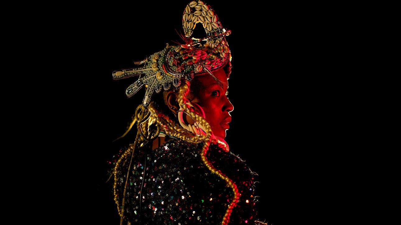 ALKANTARA - Close-up profile portrait of a Black woman illuminated by a warm red theatrical spotlight against a black background. She wears an elaborate headpiece decorated with cowrie shells and metallic details, long blonde braids, and a sparkling sequined outfit. - ©Carolina Maya