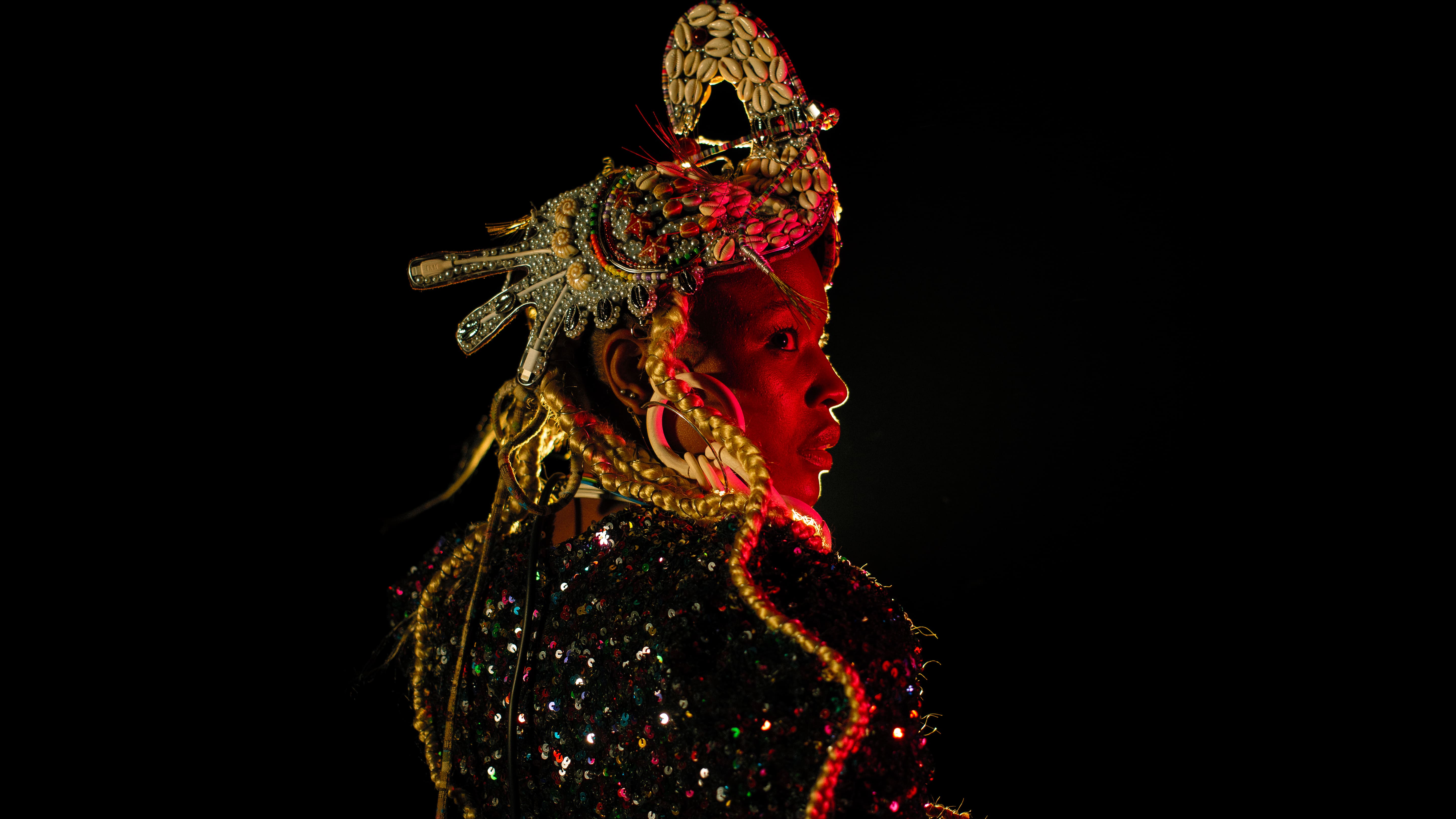 Close-up profile portrait of a Black woman illuminated by a warm red theatrical spotlight against a black background. She wears an elaborate headpiece decorated with cowrie shells and metallic details, long blonde braids, and a sparkling sequined outfit. - Carolina Maya