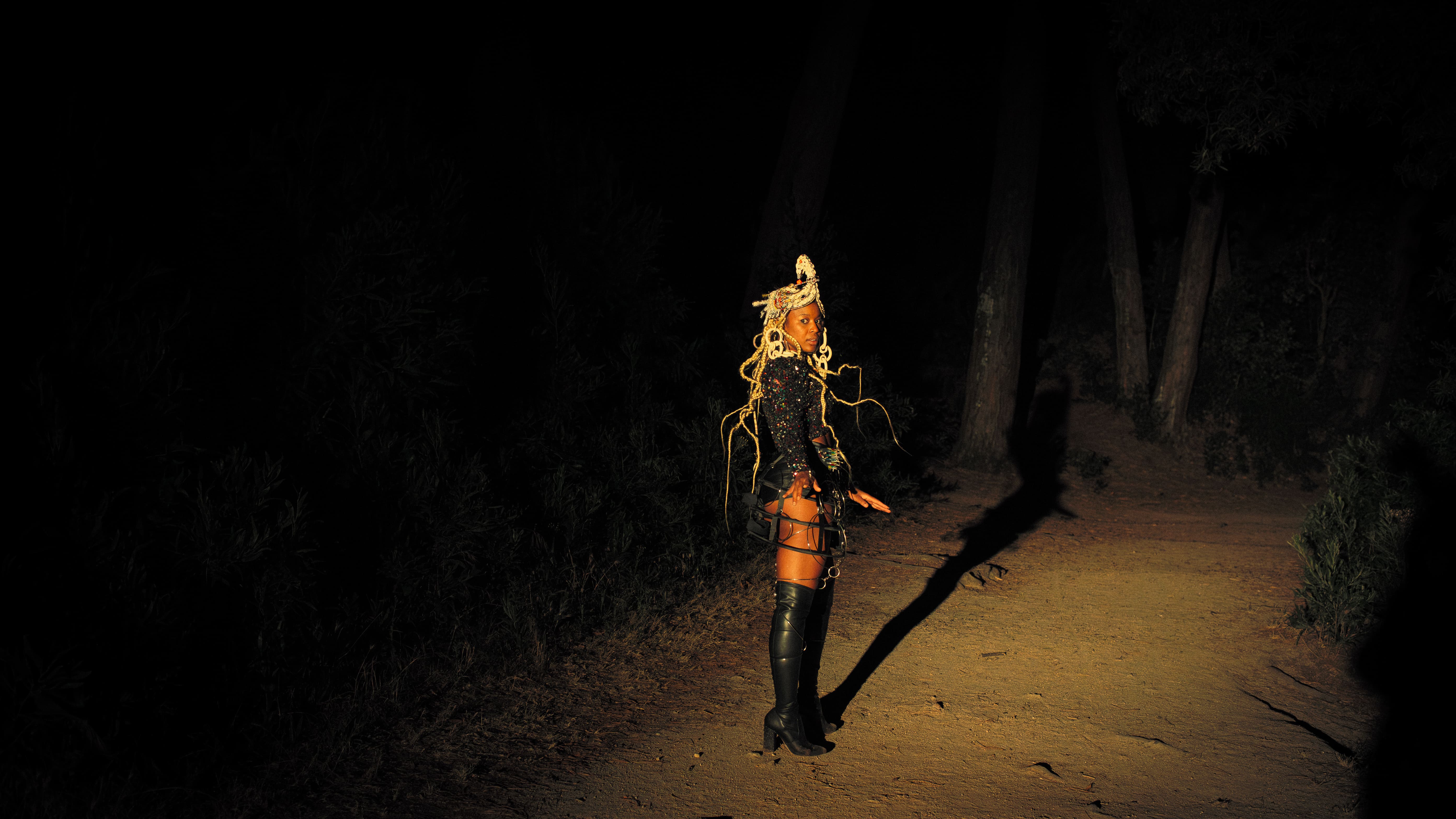 Full-length shot of a Black woman standing on a dirt path at night, surrounded by the darkness of a forest. Illuminated by a yellow spotlight, she wears a shimmering sequined top, thigh-high black boots, a cowrie-shell headpiece, and long loose braids. - Carolina Maya