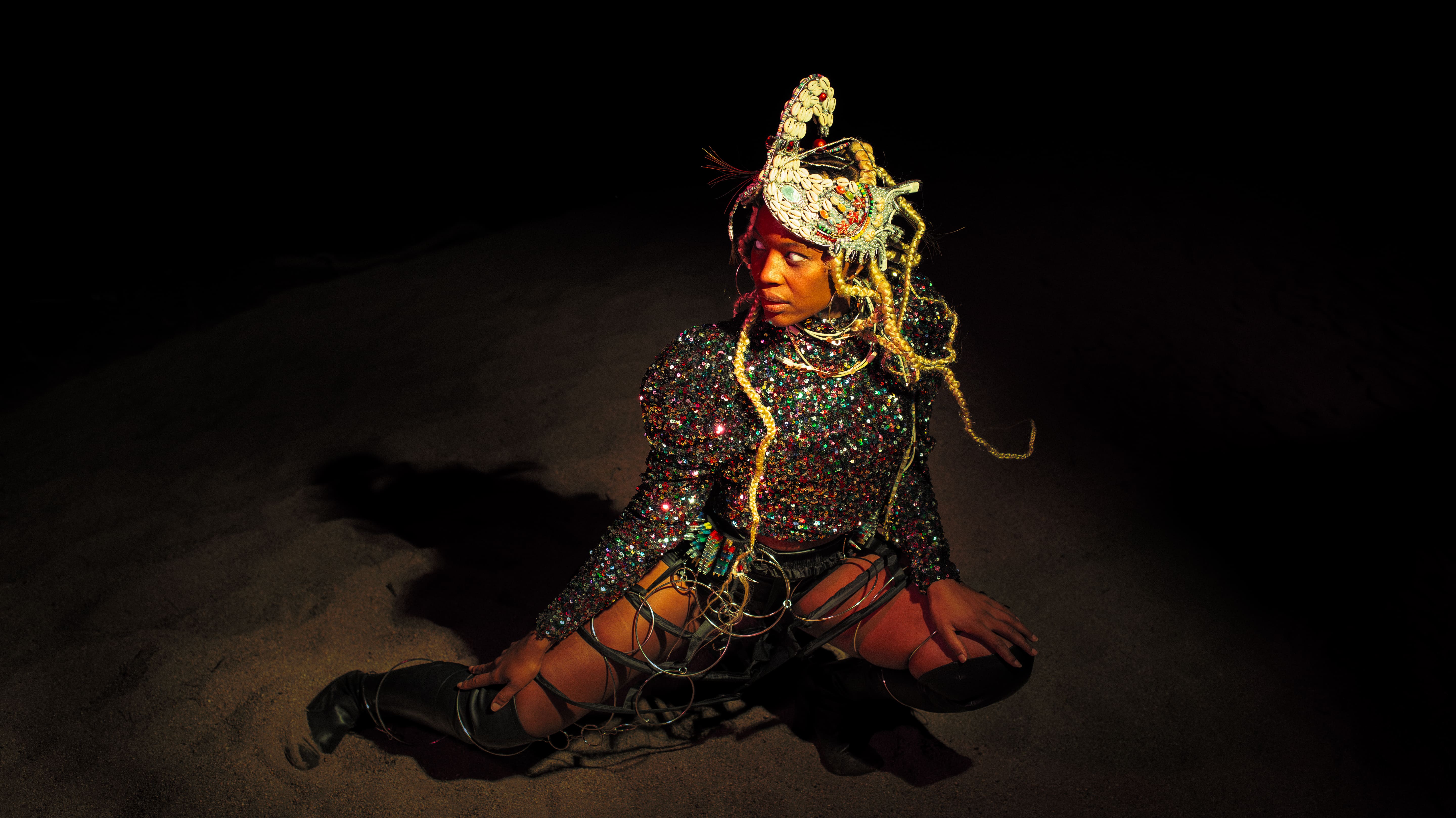 Medium shot of a Black woman kneeling in the sand in pitch darkness under a warm spotlight. She wears a glittering sequined top, high black boots, a metallic cage skirt structure, and a detailed headpiece with cowrie shells and blonde braids. - Carolina Maya